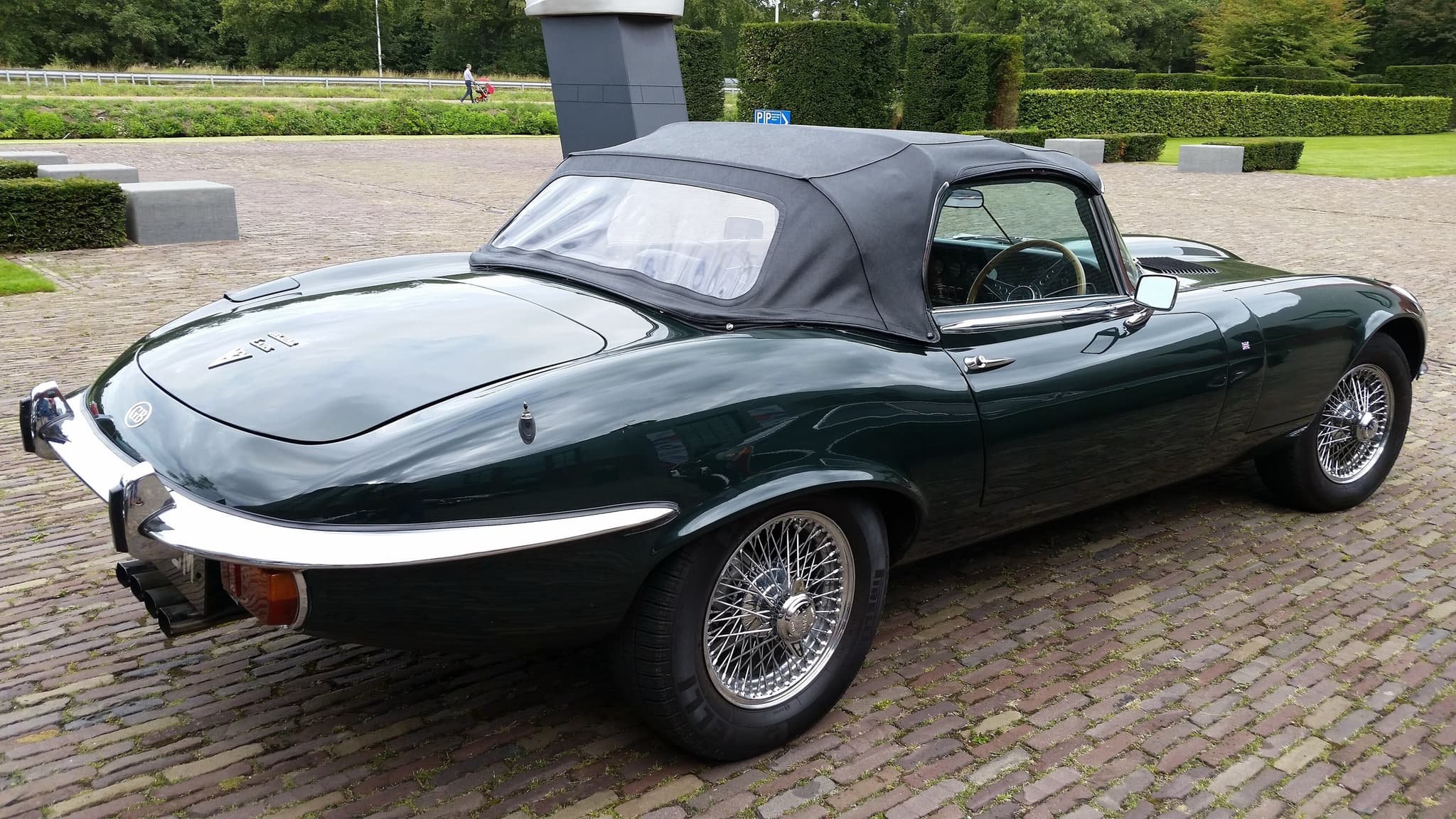 A dark green E-Type roadster with its hood up from behind on cobbles, showing a 'V12' badge and four exhaust pipes. Opens a larger view