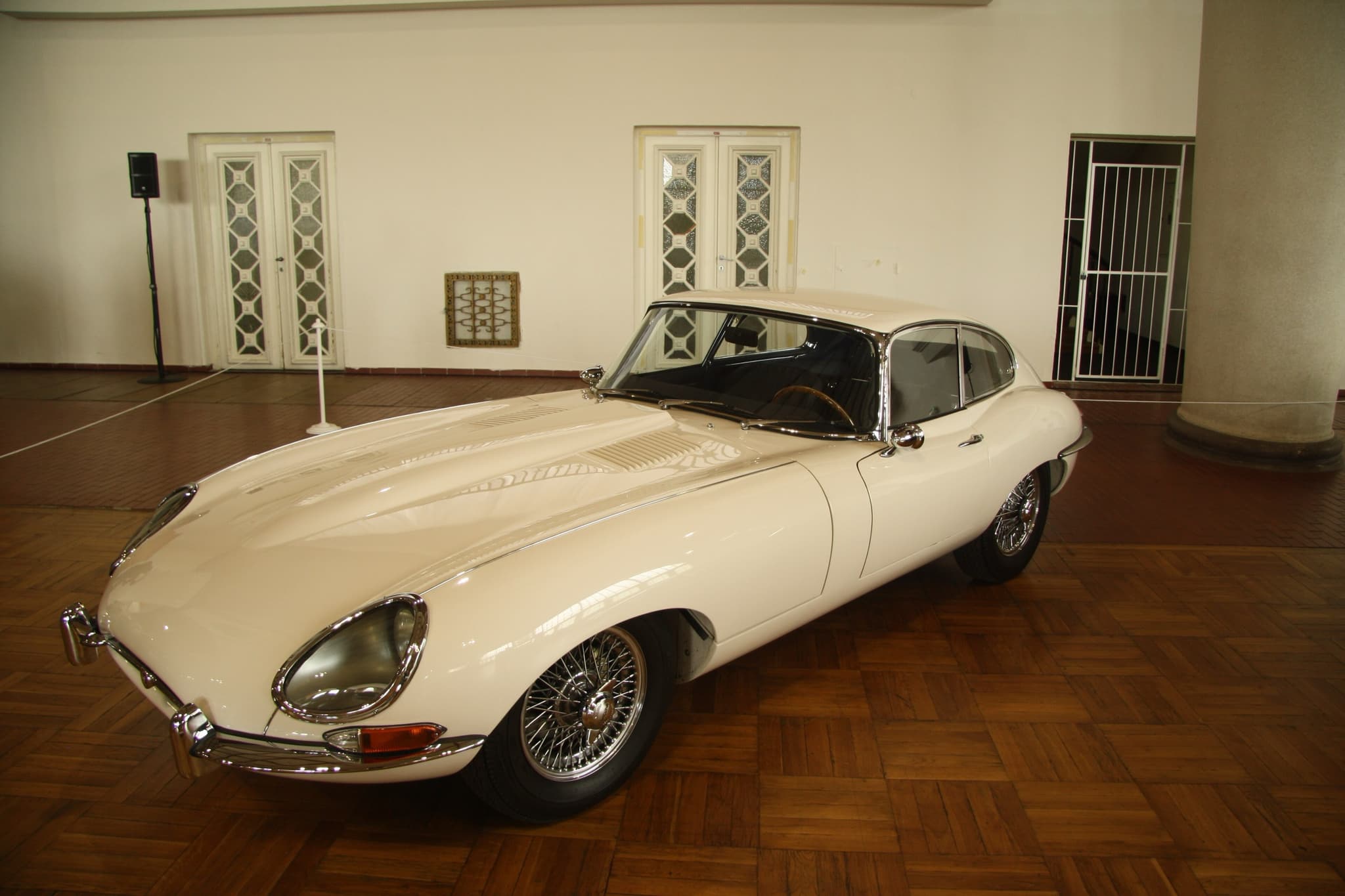 A white E-Type coupé on a parquet floor in a tall white hall with patterned glass doors.