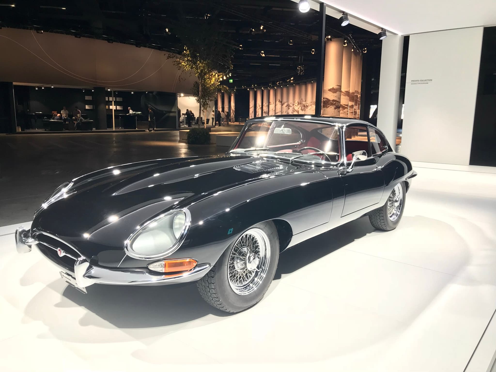 A black E-Type coupé with red leather seats on a white display floor, under exhibition spotlights.