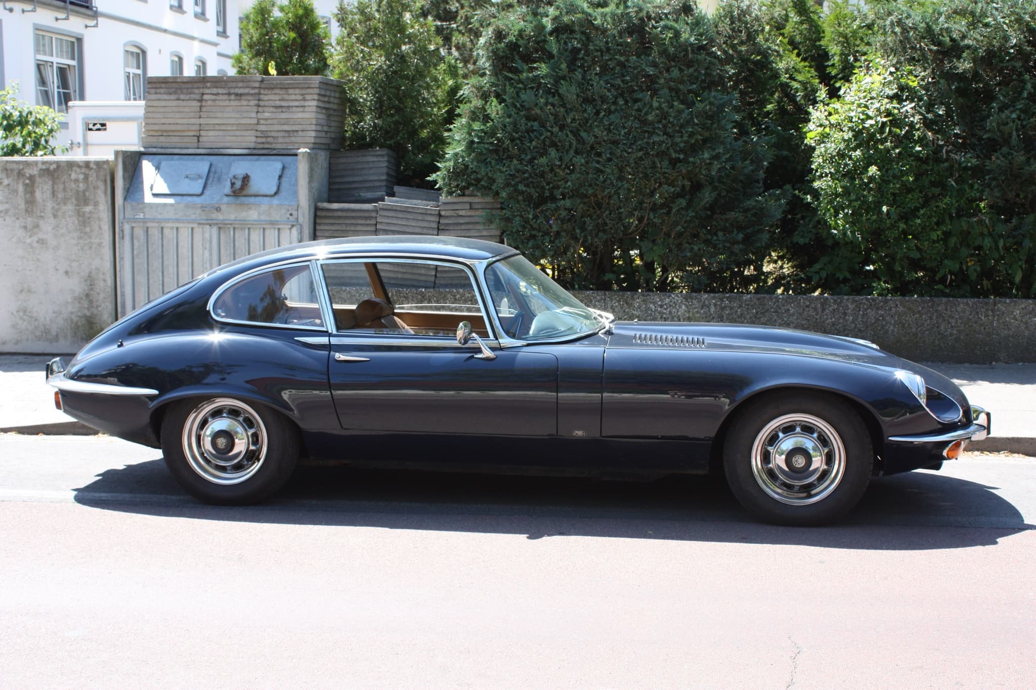 A dark blue E-Type 2+2 with flared wheel arches and a long roof, in profile at the kerb by a hedge.