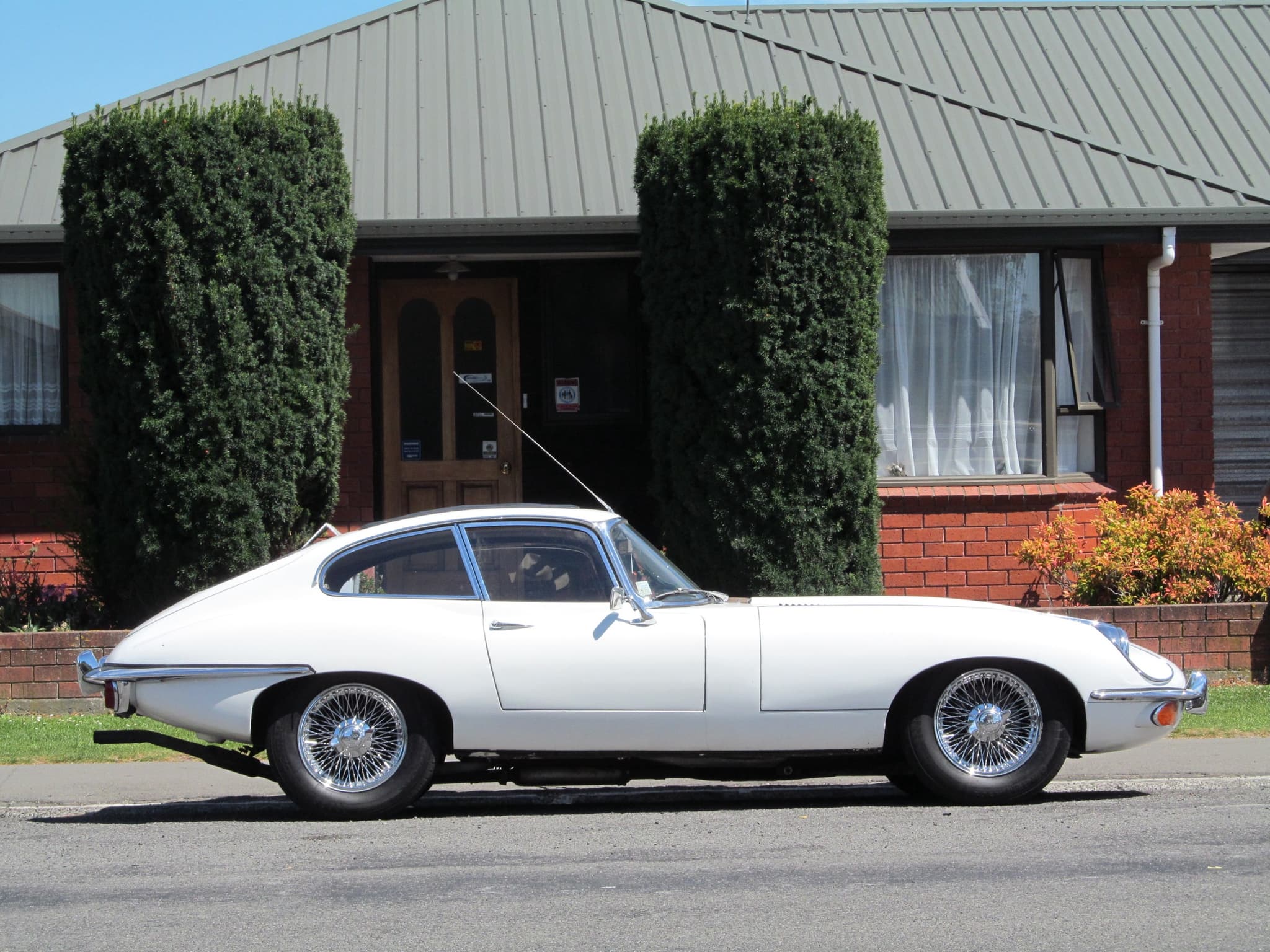 A white E-Type coupé in profile at the kerb of a quiet suburban street, in front of a brick bungalow. Opens a larger view