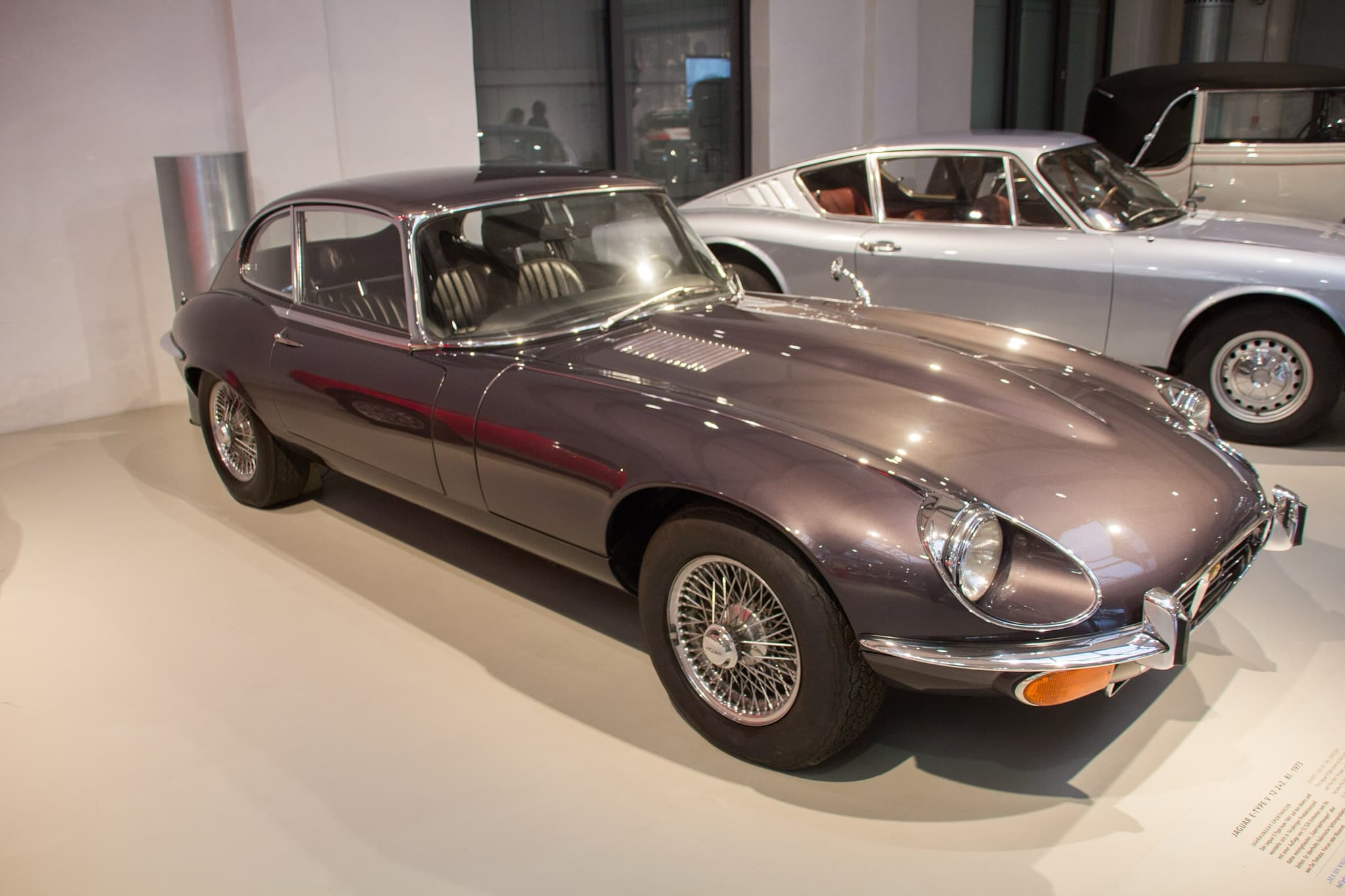 A dark grey E-Type 2+2 with a cross-hatched grille and wire wheels on a pale museum floor. Opens a larger view