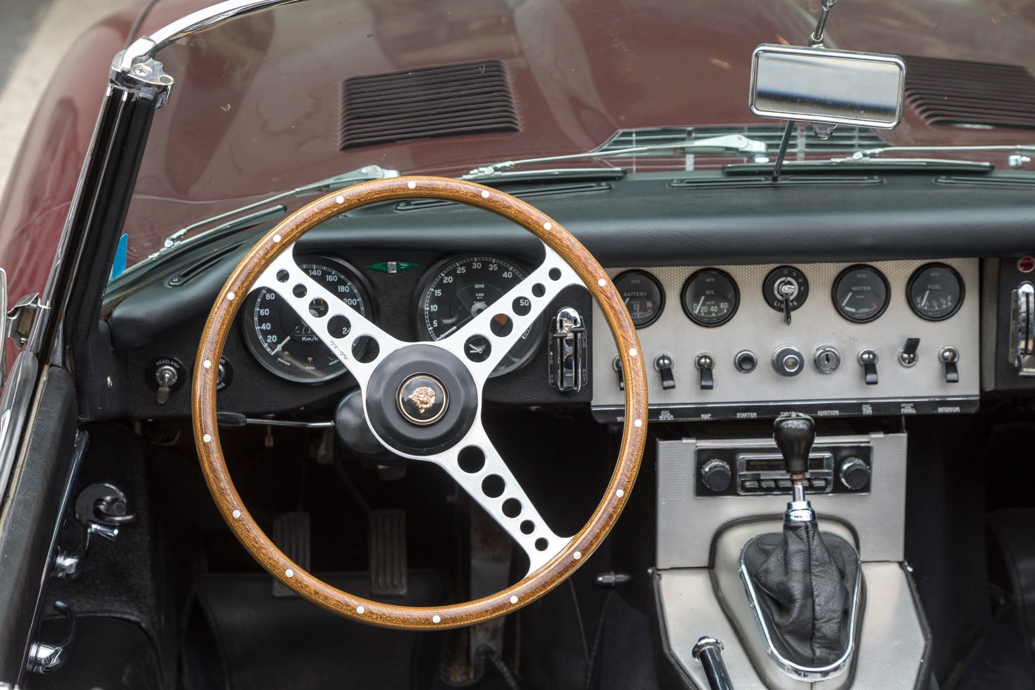 The dash of an early E-Type roadster: a riveted wood-rim wheel on the left and an aluminium centre panel with dials and toggle switches.