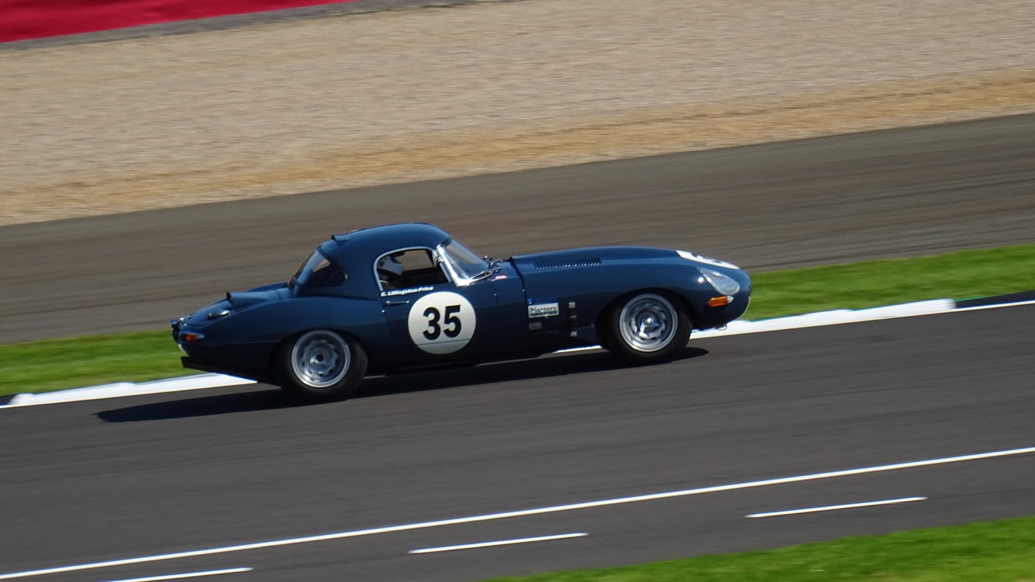 A dark blue E-Type roadster with a hardtop, numbered 35, racing on a circuit beside a strip of grass. Opens a larger view