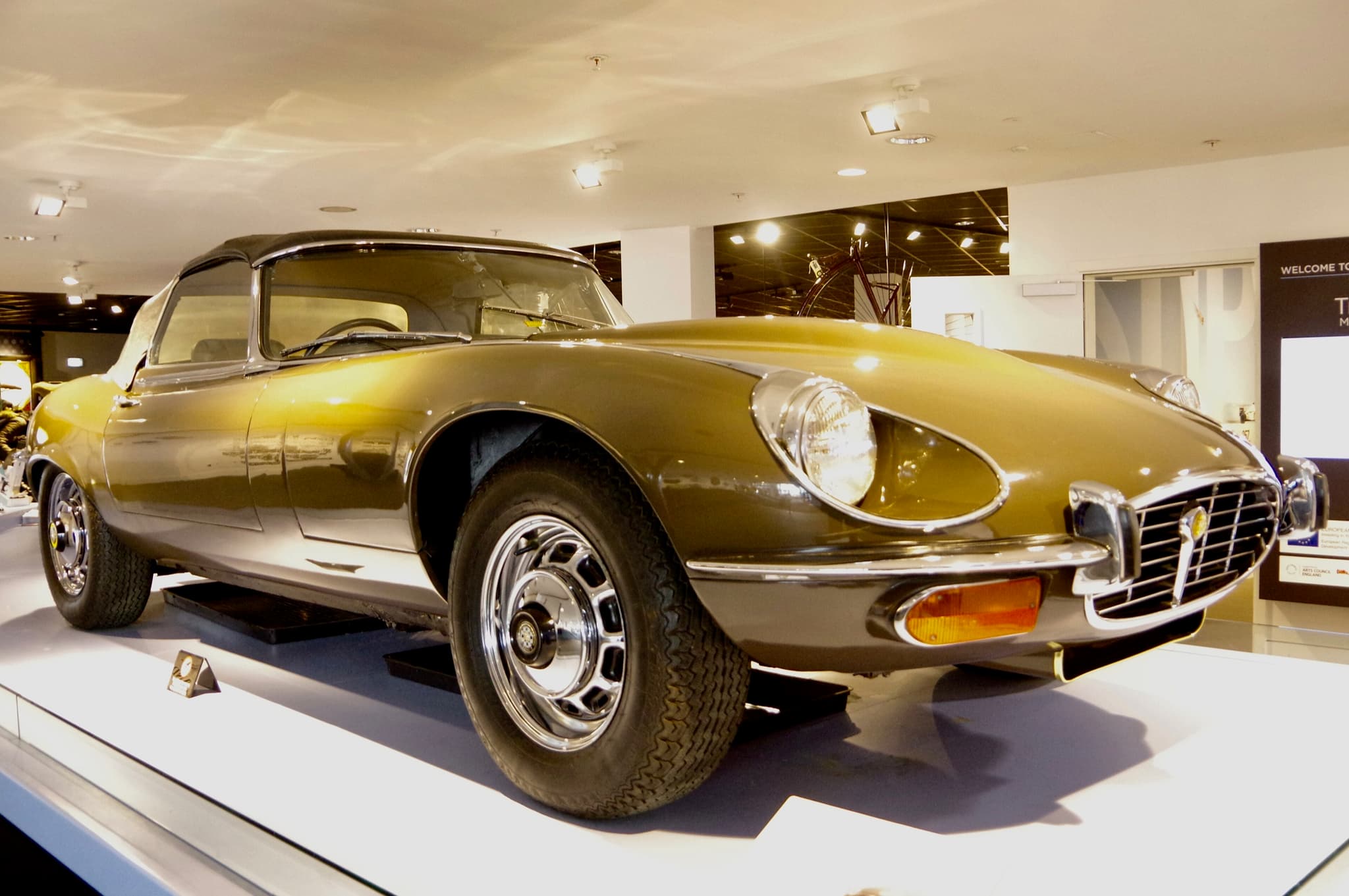 A bronze-green E-Type roadster with its hood up and a cross-hatched grille on a white museum plinth.