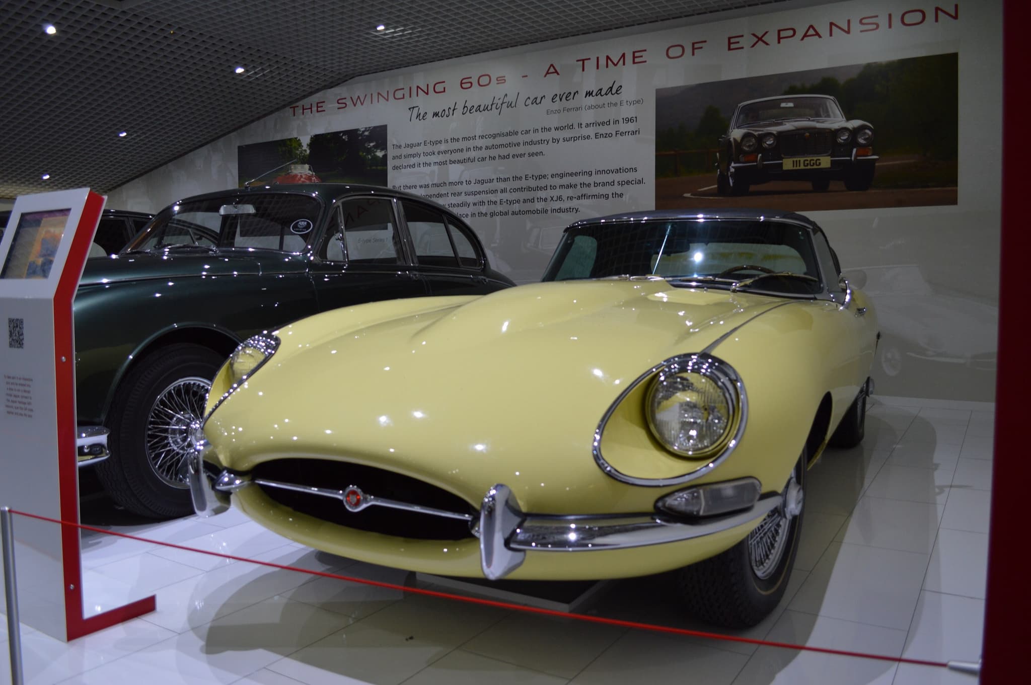 A pale yellow E-Type coupé in a museum display beneath a wall panel headed 'The Swinging 60s'.