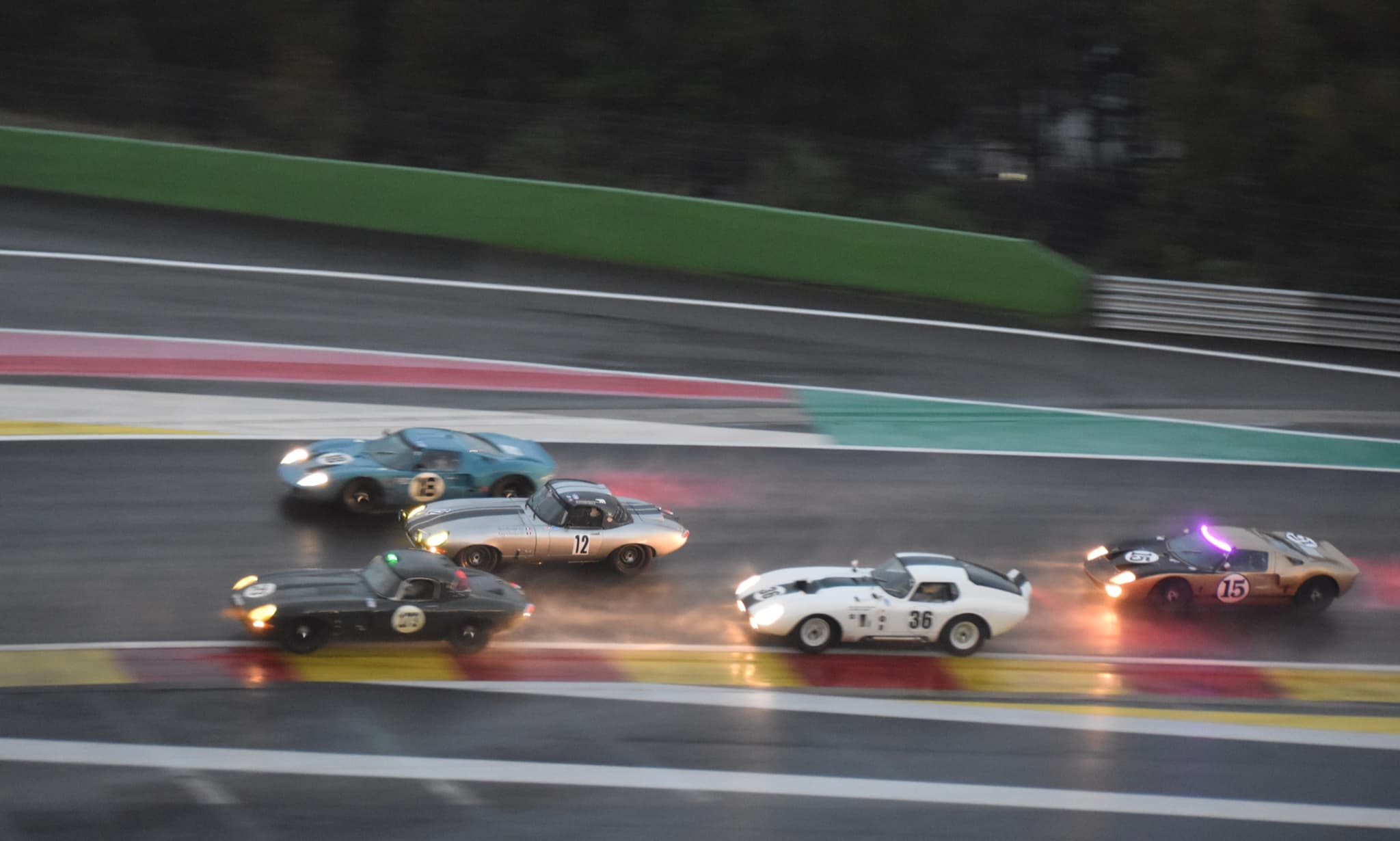 Six racing sports cars of the 1960s, two of them E-Types, on a wet track at dusk with headlamps on.