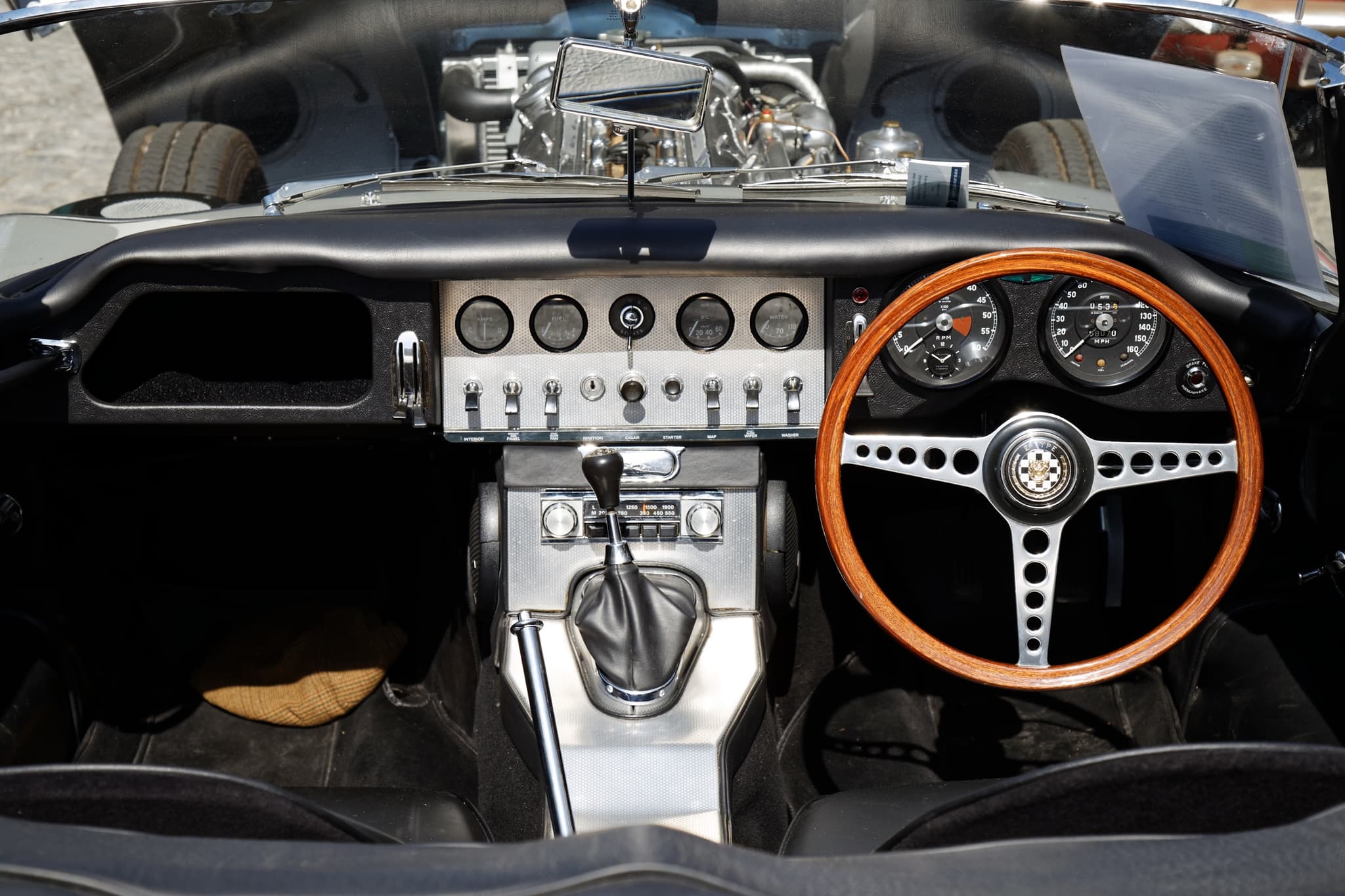 The cockpit of an early E-Type roadster: an aluminium centre panel with five dials and a row of toggle switches beside a wood-rim wheel.