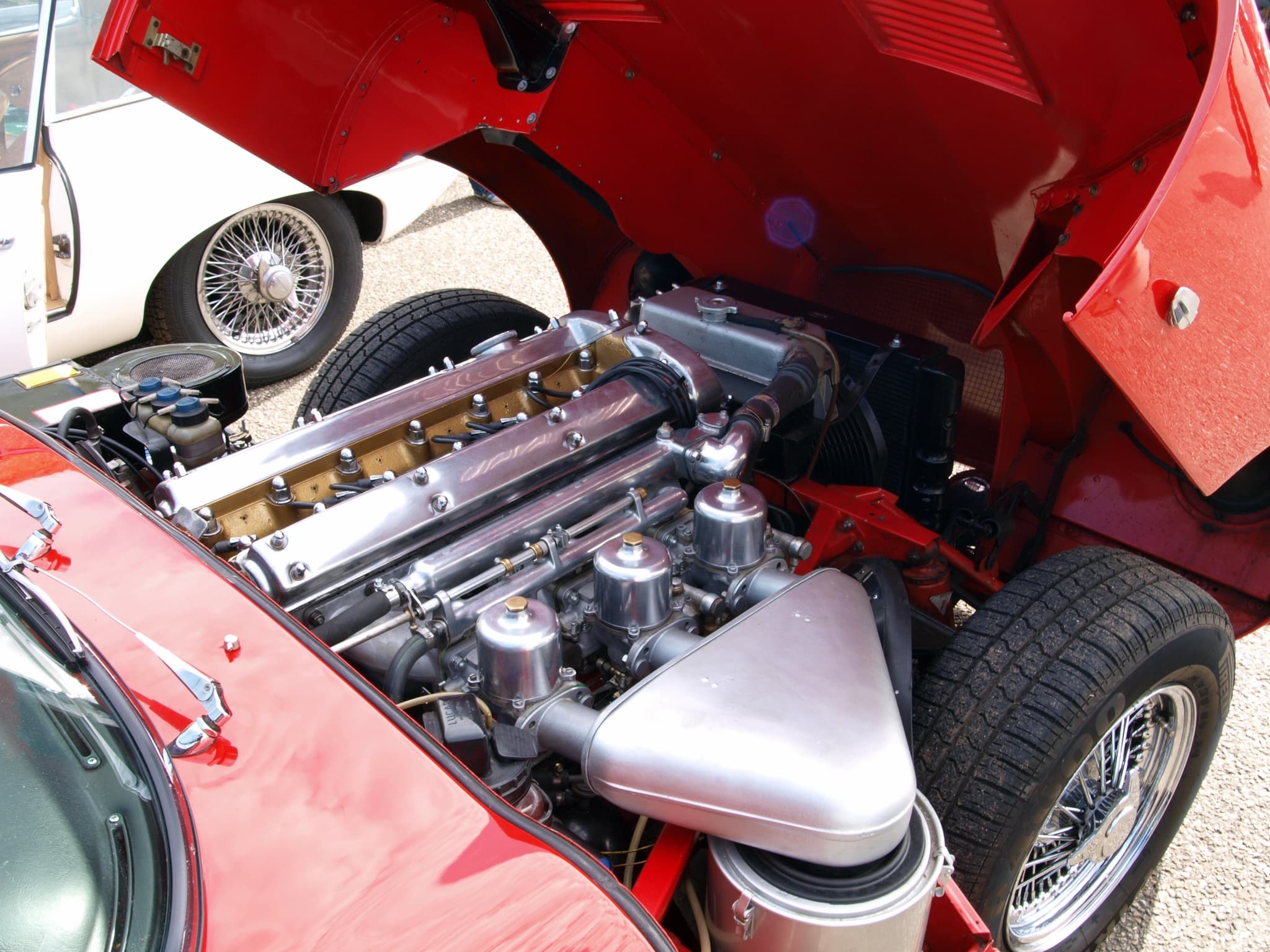 The engine of a red E-Type roadster with its bonnet tipped forward: polished cam covers, three carburettors and a round air filter.