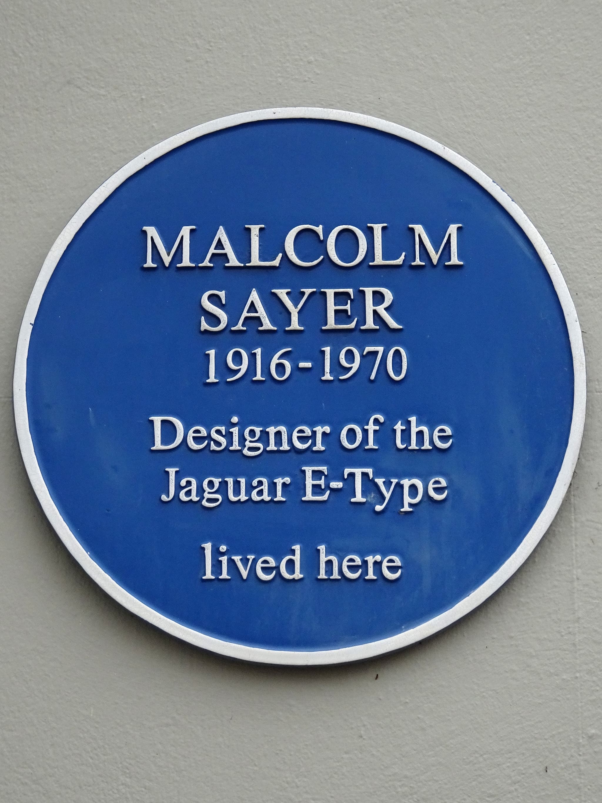 A round blue plaque reading Malcolm Sayer 1916-1970, Designer of the Jaguar E-Type, lived here, on a grey wall.