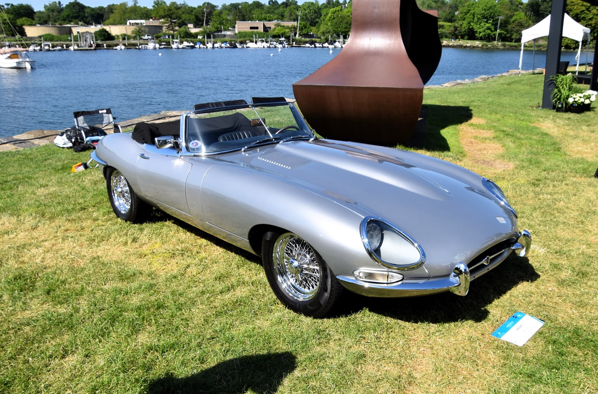 A silver E-Type roadster with its hood folded and glass-covered headlamps, parked on grass by a harbour full of moored boats. Opens a larger view