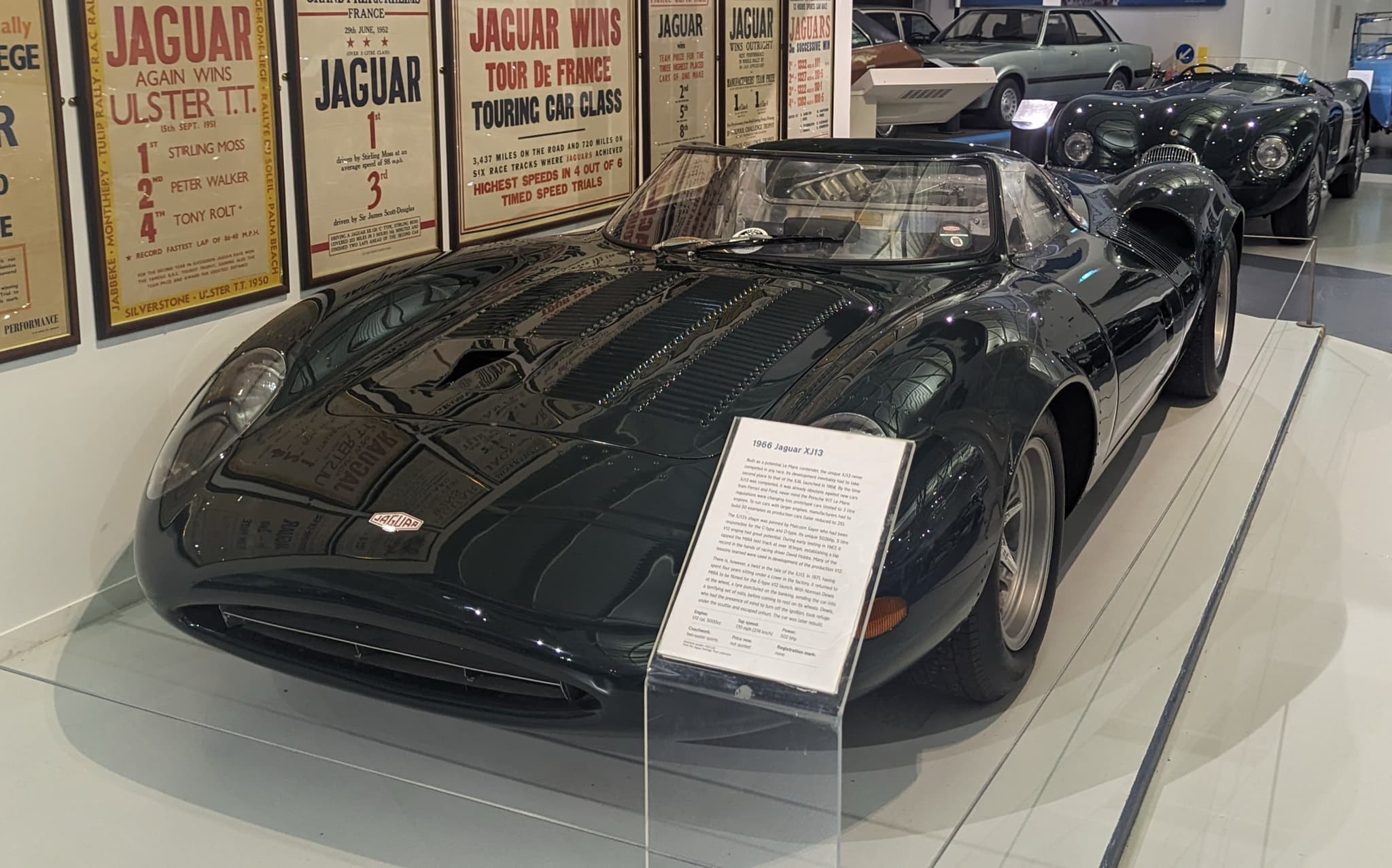 A dark green mid-engined Jaguar racing car behind a display rail with an information panel. Opens a larger view