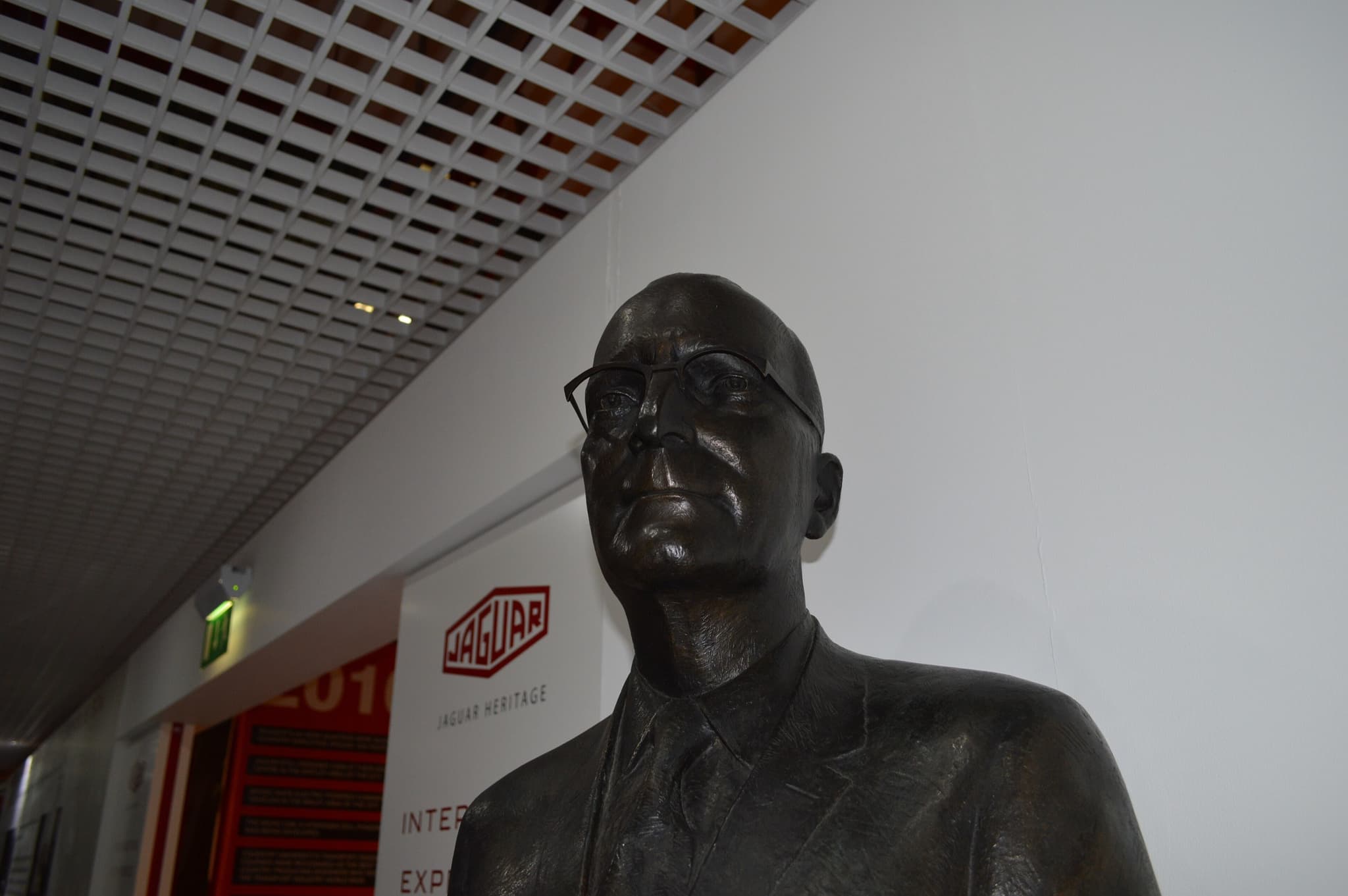 A bronze bust of a bald man in spectacles and a suit beside a Jaguar Heritage sign. Opens a larger view