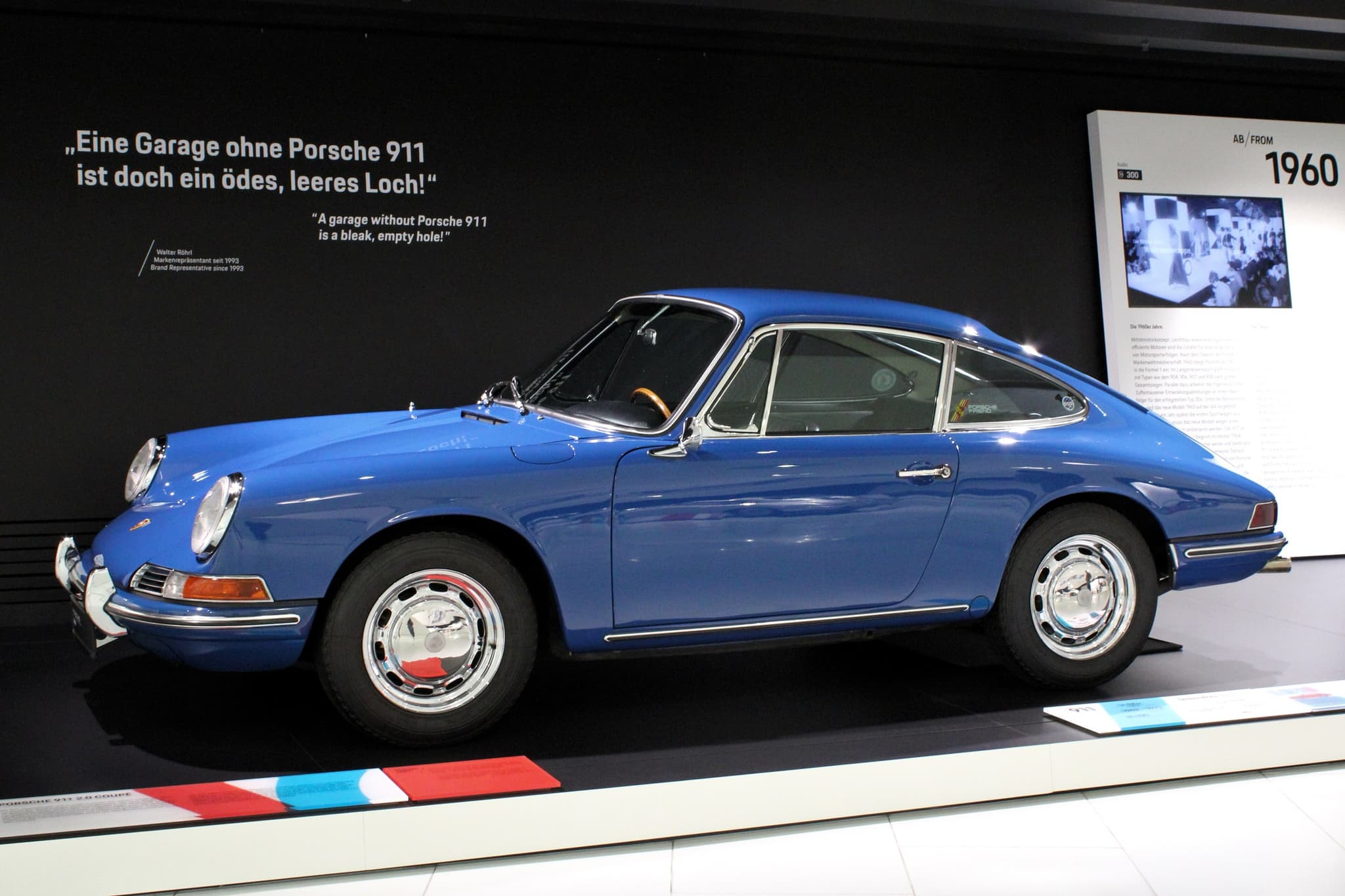 An early blue Porsche 911 coupé in profile on a museum plinth.