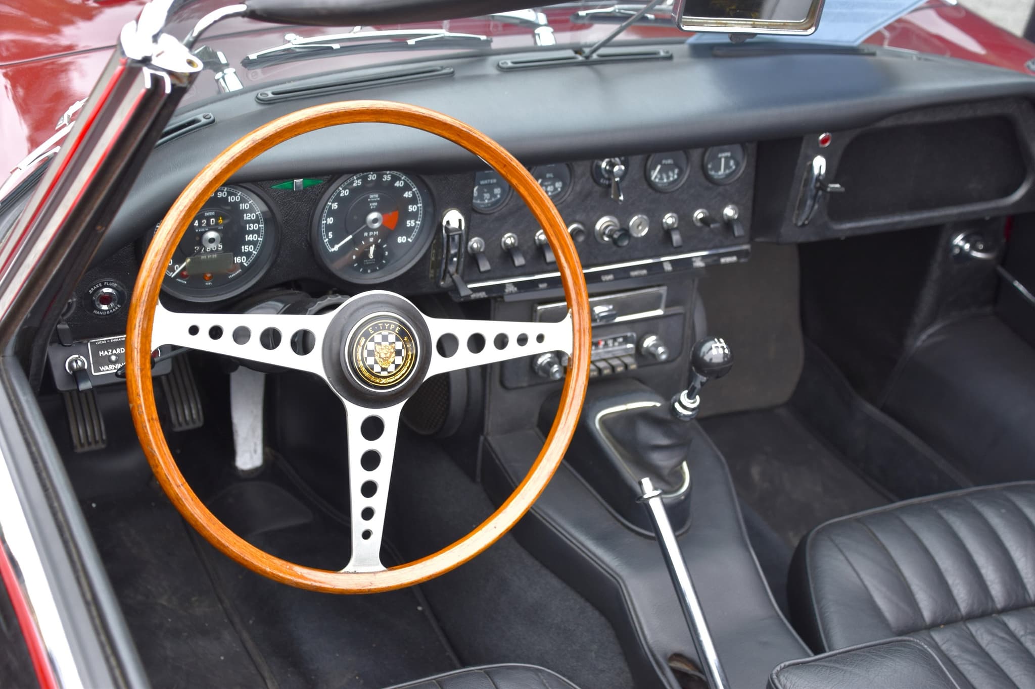 The dash of an E-Type roadster: speedometer and rev counter behind a wood-rim wheel, and a black centre panel of dials and toggle switches.
