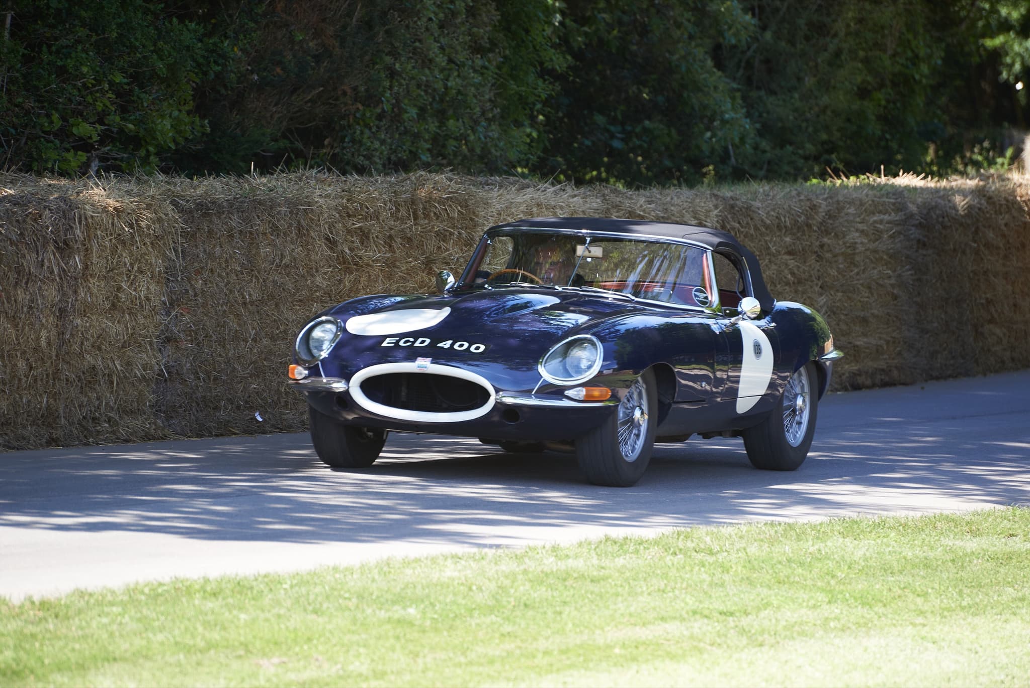 A dark blue E-Type roadster with a hardtop driving past straw bales under trees.