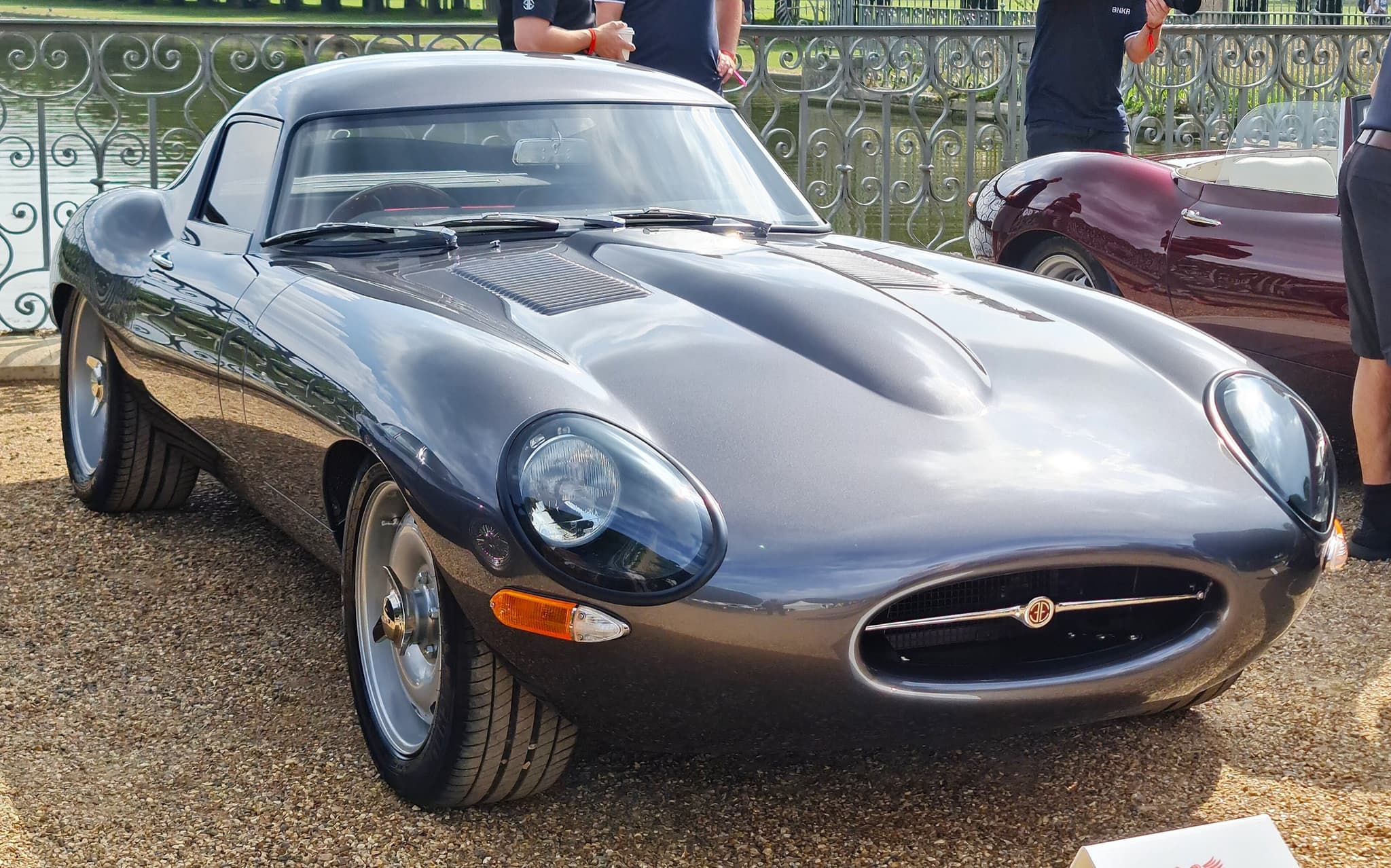 A dark grey fastback recreation of the E-Type Low Drag Coupé, without bumpers, on gravel by a railing.