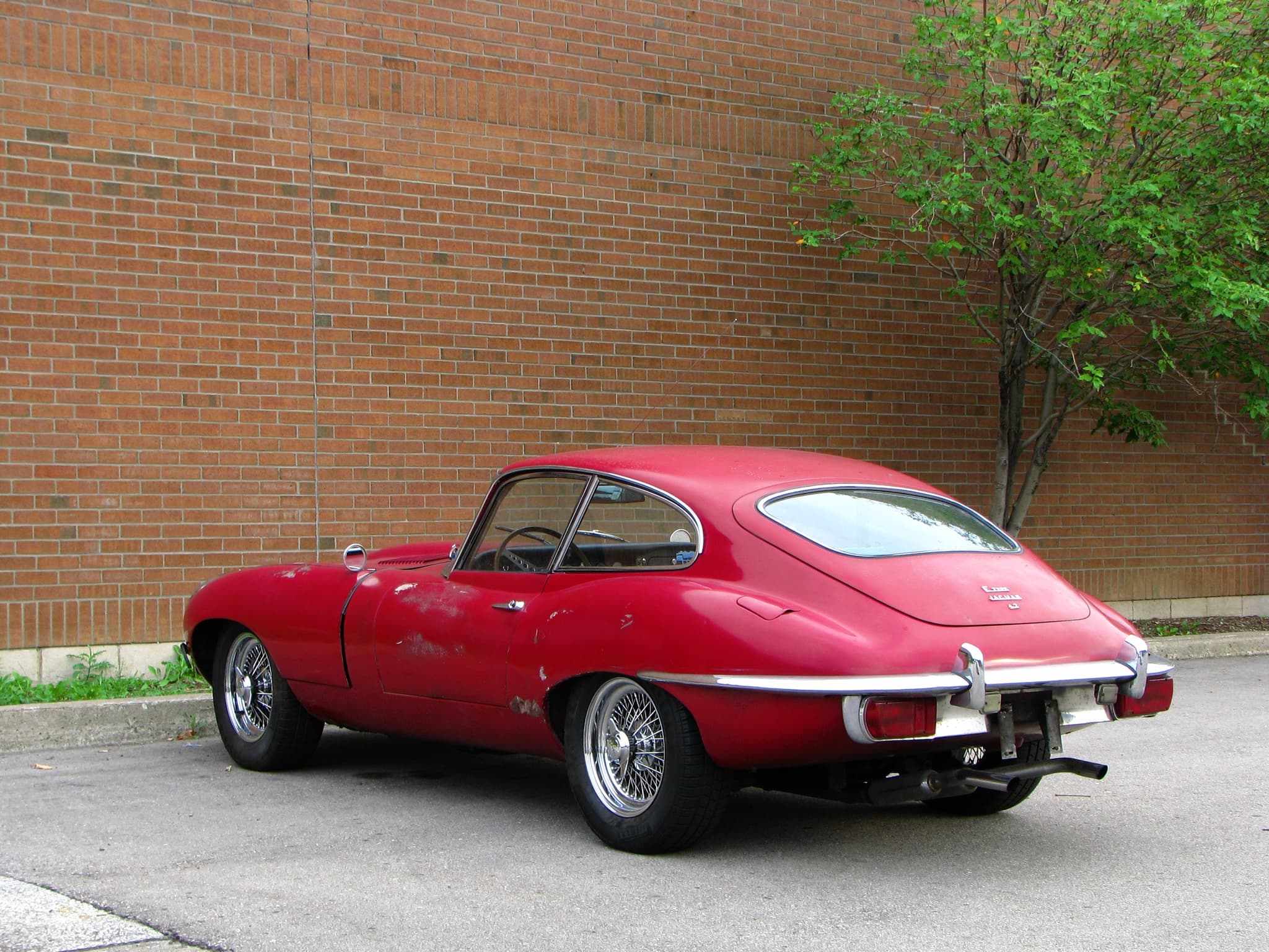 A faded red E-Type coupé from behind in a car park by a brick wall, with large tail lamps below the bumper.