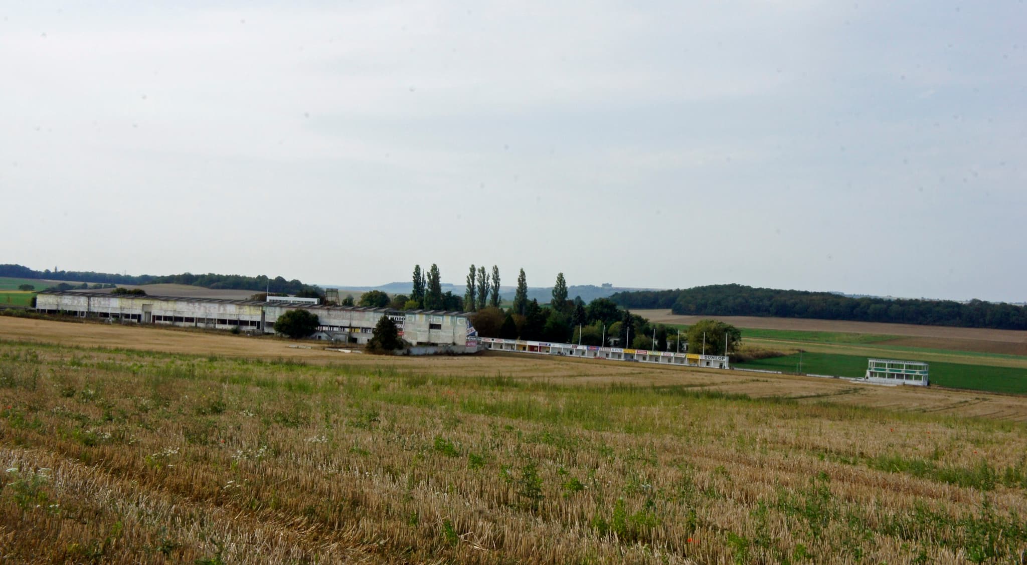 Old grandstands and pit buildings beside a road beyond a stubble field. Opens a larger view