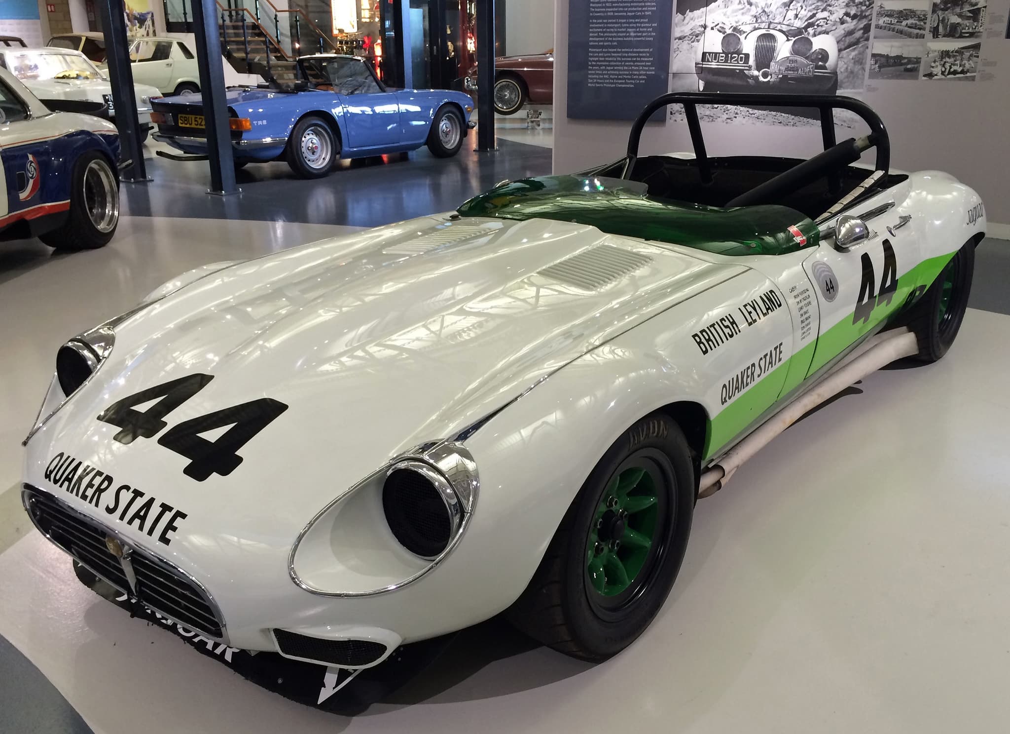A white E-Type V12 roadster with a green stripe, a roll hoop and the number 44 in a museum.