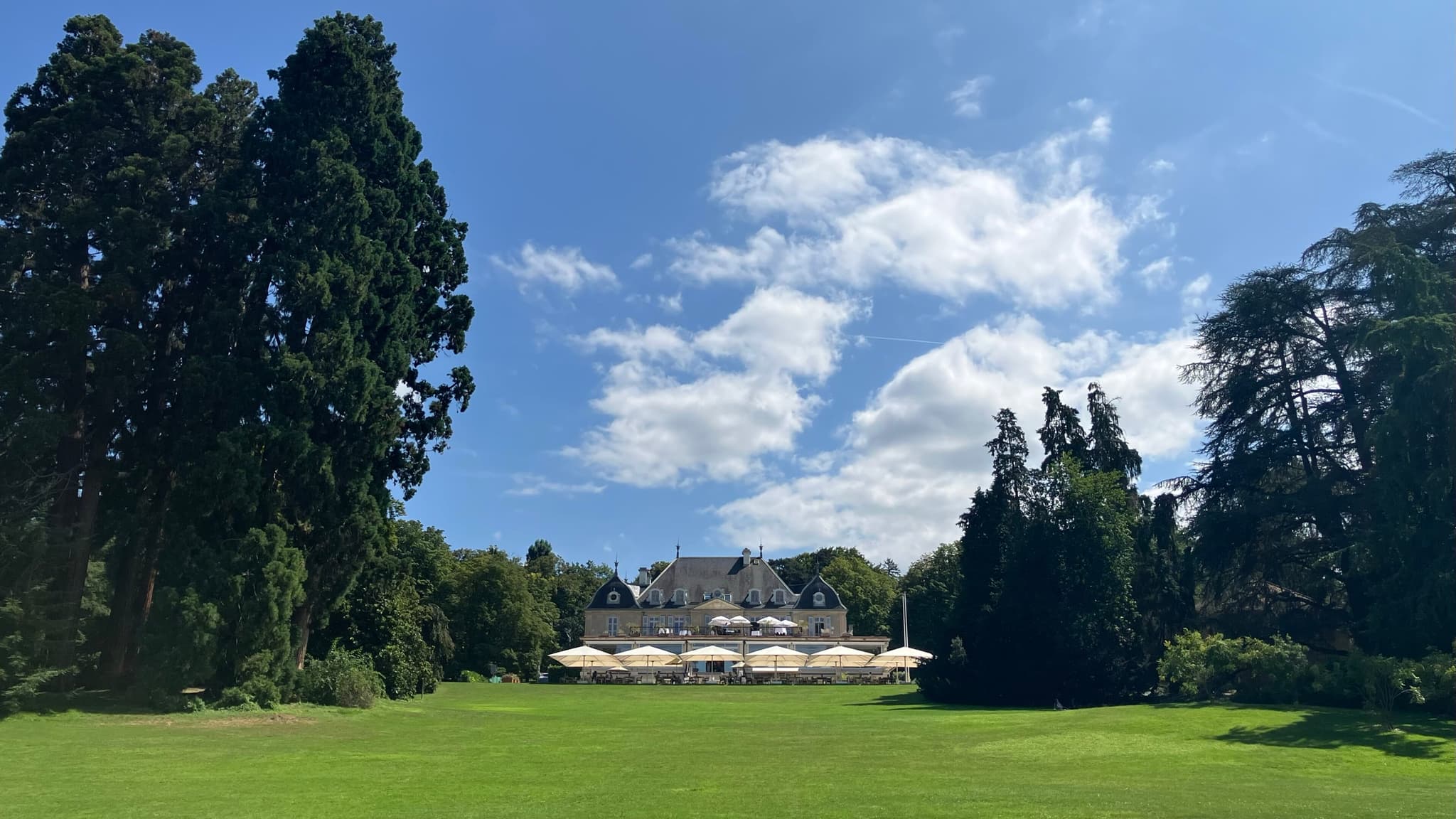 A wide lawn leading to a restaurant building with white parasols among tall cedars. Opens a larger view