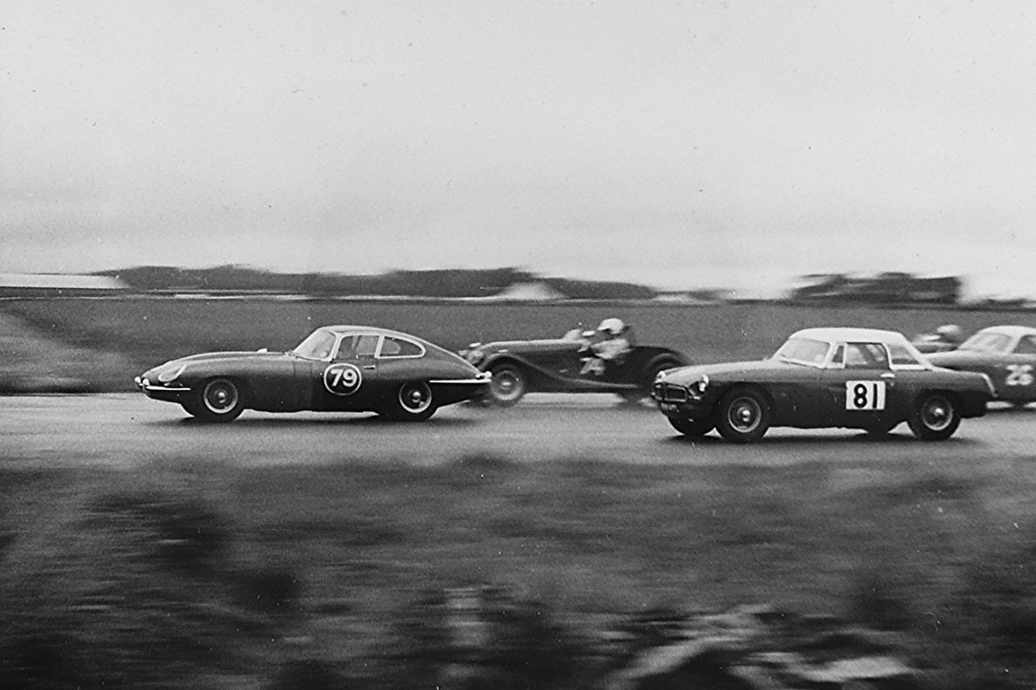 A black-and-white photograph of an E-Type coupé numbered 79 racing alongside a Morgan and an MGB. Opens a larger view