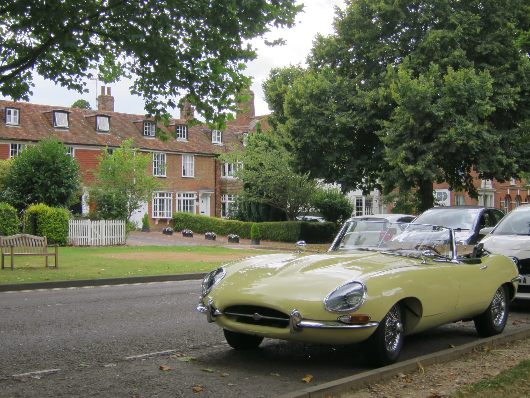 A pale green E-Type roadster parked beside a village green with a row of brick houses.