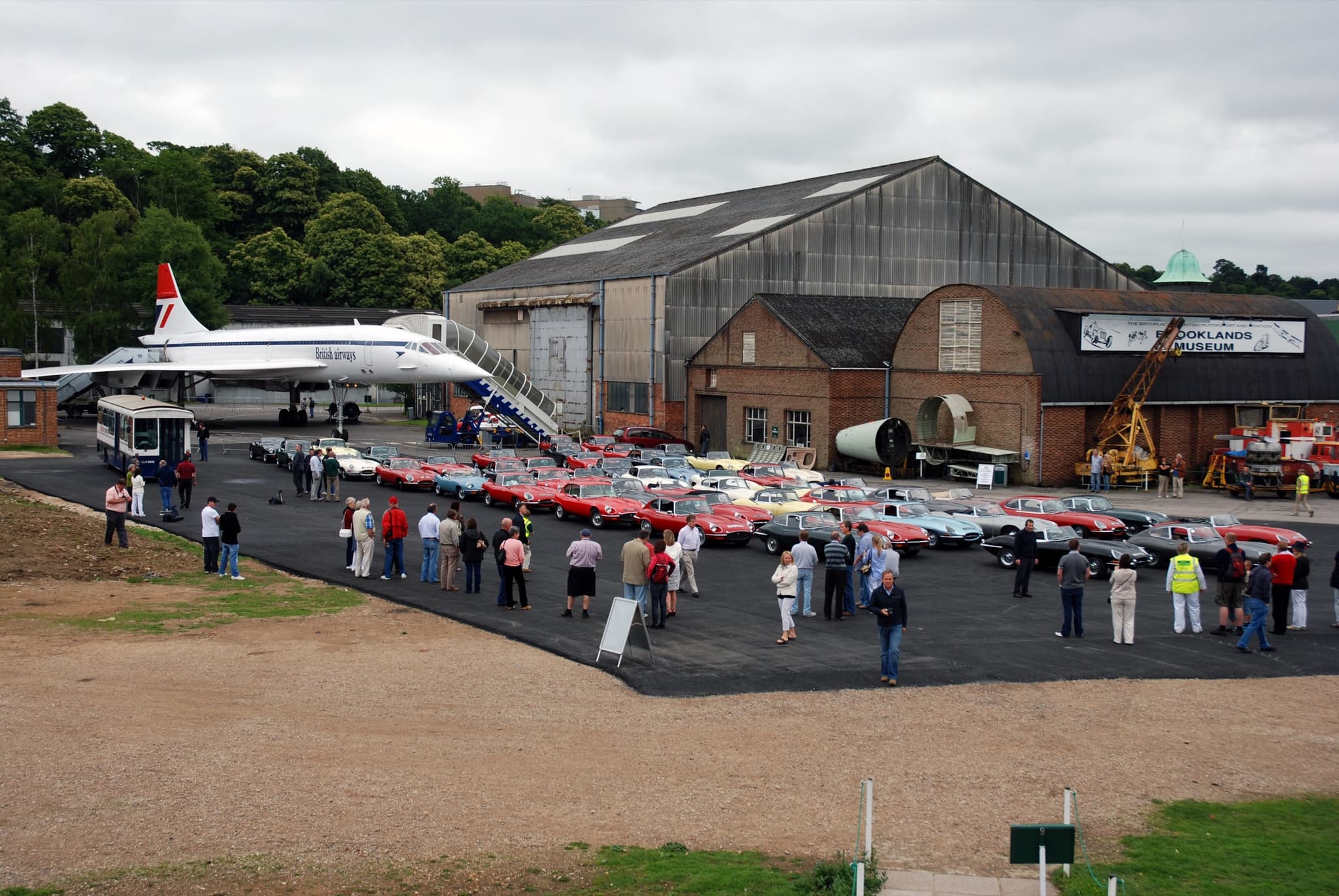 Rows of E-Types parked on an apron at Brooklands, with Concorde and museum hangars behind.