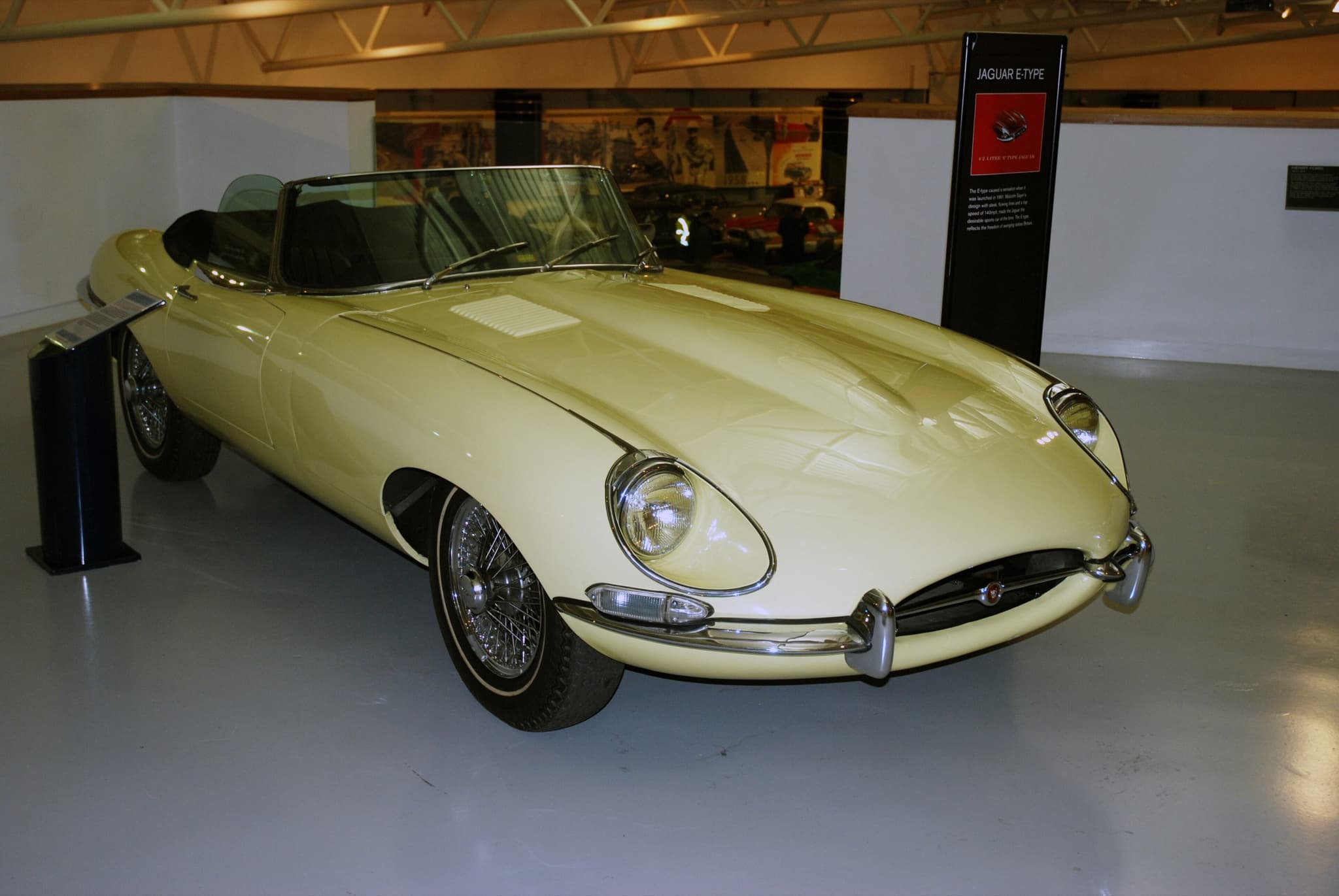 A pale yellow E-Type roadster with open headlamps and whitewall tyres, displayed in a museum hall.