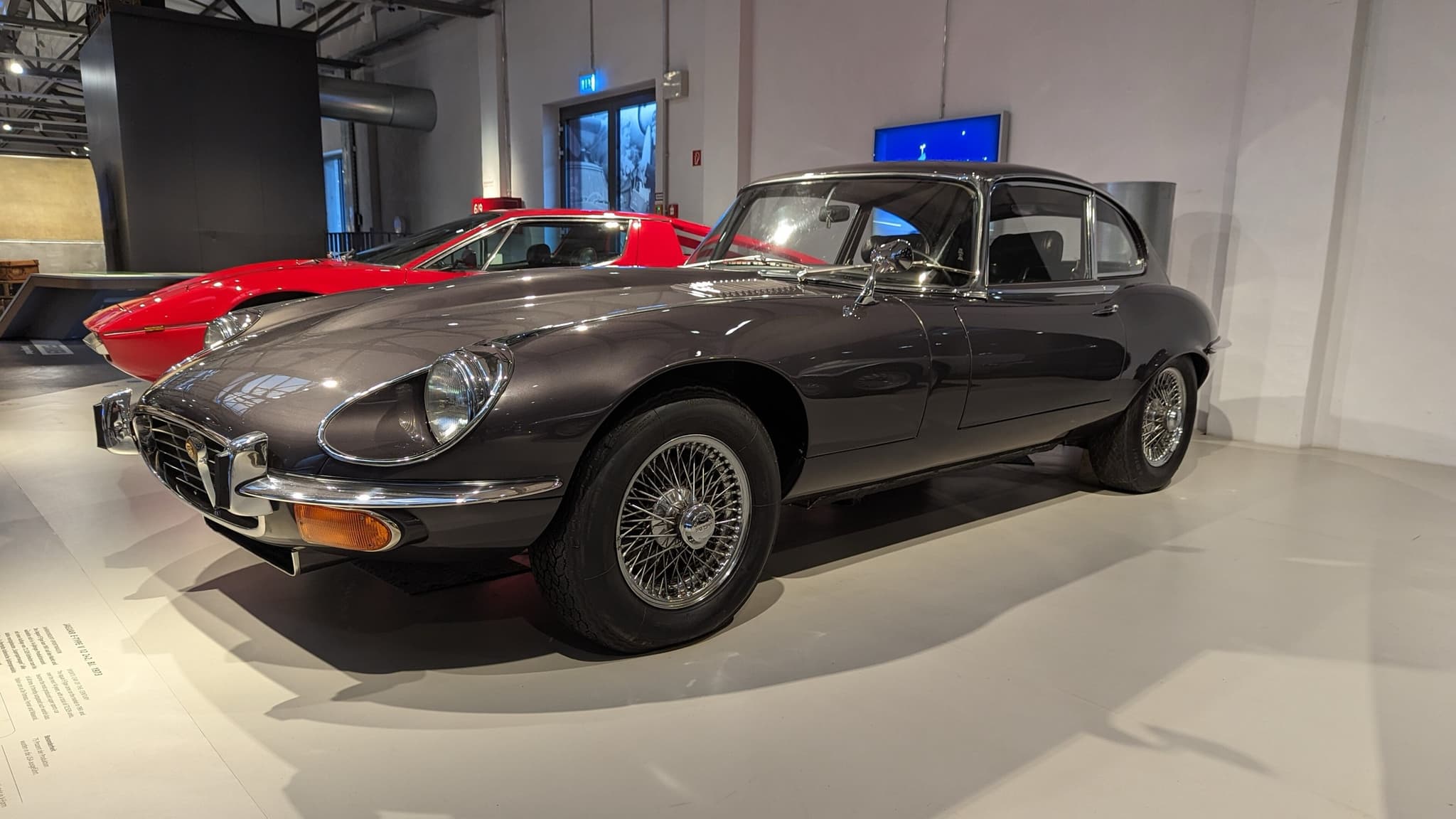 A grey E-Type 2+2 with a cross-hatched grille and flared wheel arches in a museum hall.