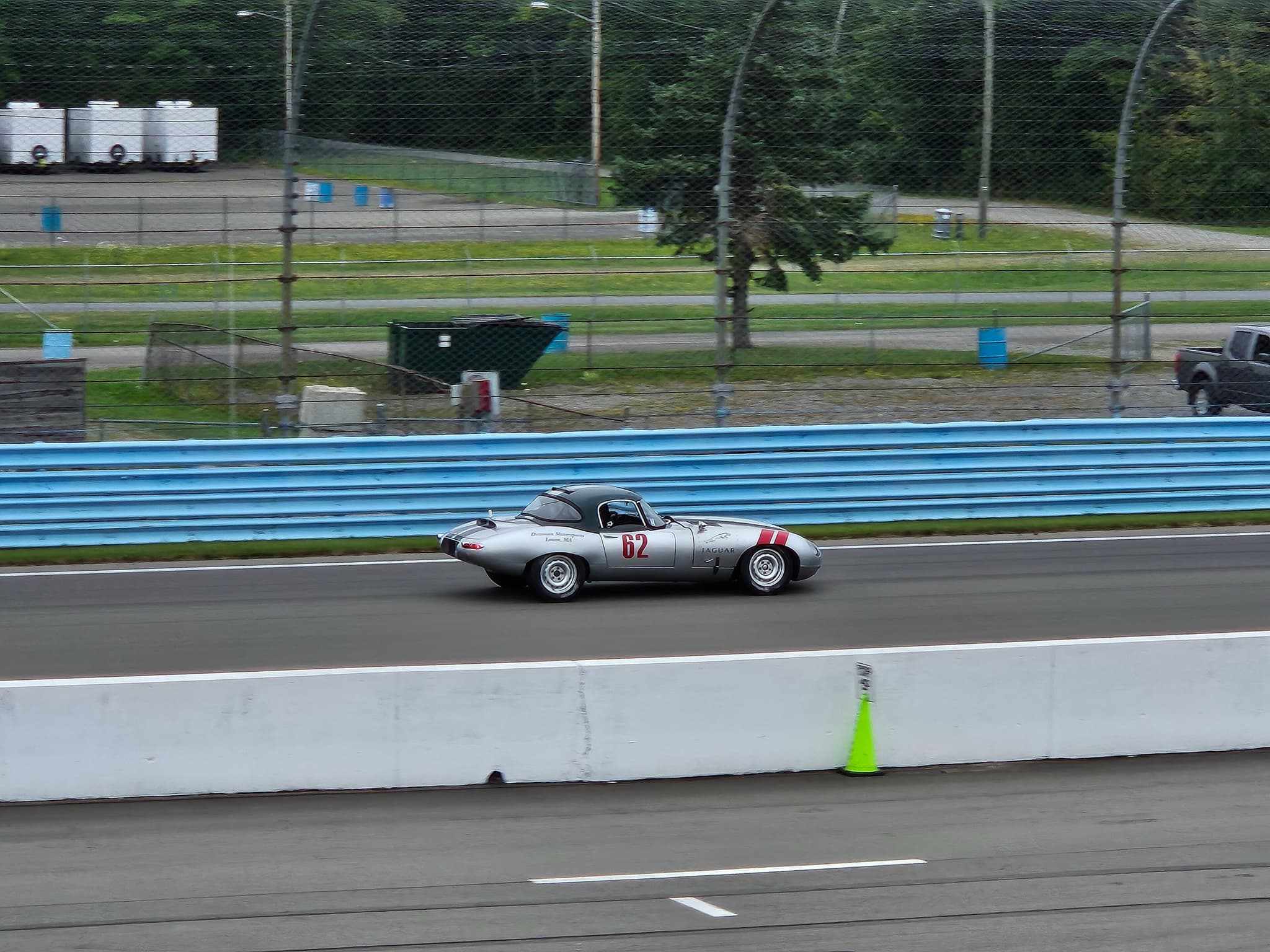 A silver E-Type coupé with red stripes racing on a circuit lined with a concrete wall and fencing.