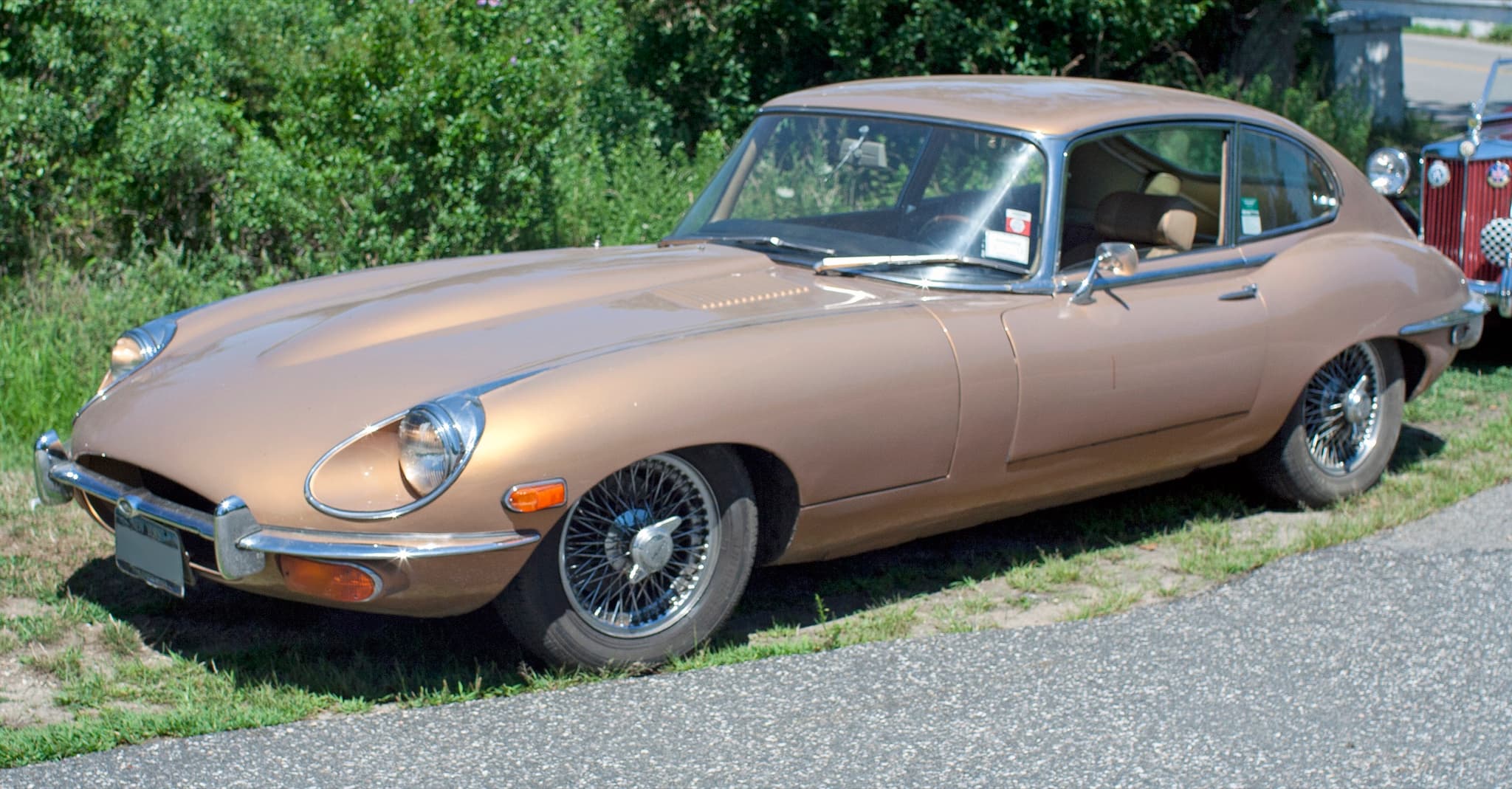 A gold E-Type 2+2 on grass by a road, with a large oval intake and the indicators below the bumper.