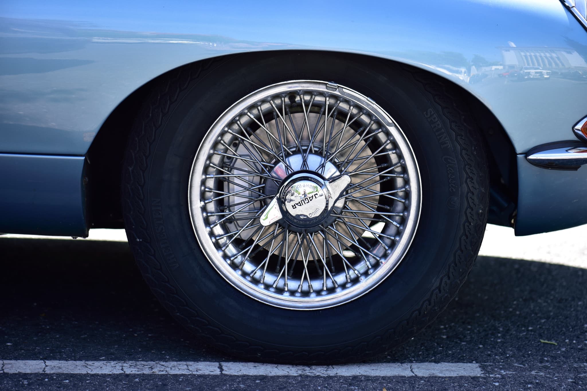 A wire wheel with a two-eared chrome knock-off spinner marked 'JAGUAR', on a pale blue E-Type. Opens a larger view