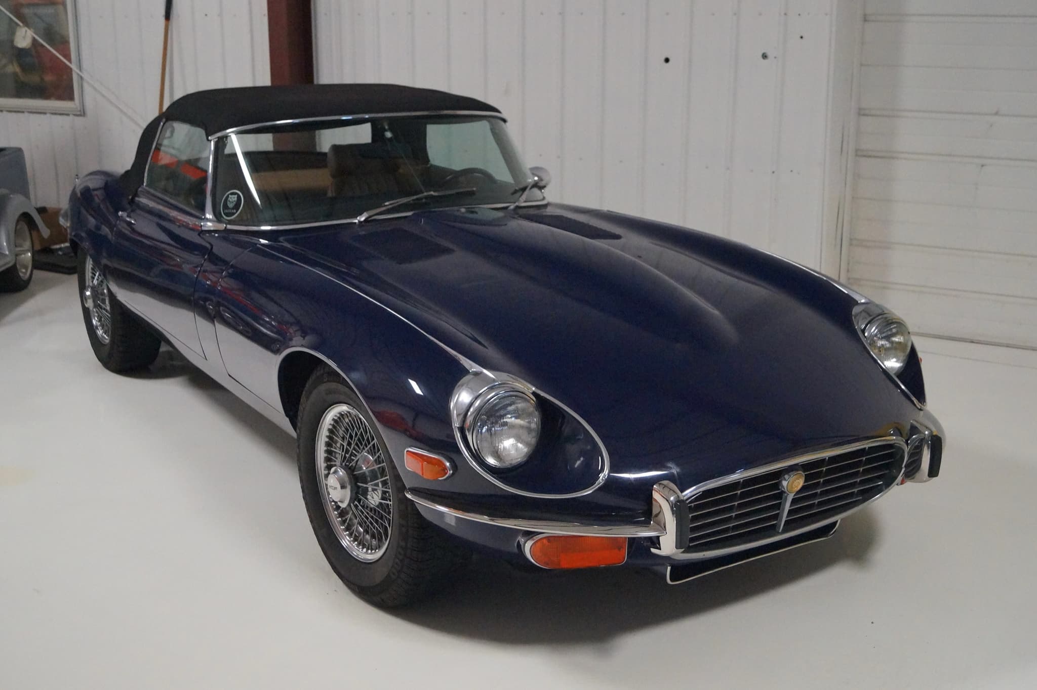 A dark blue E-Type roadster with its hood up and a cross-hatched grille in a white garage. Opens a larger view