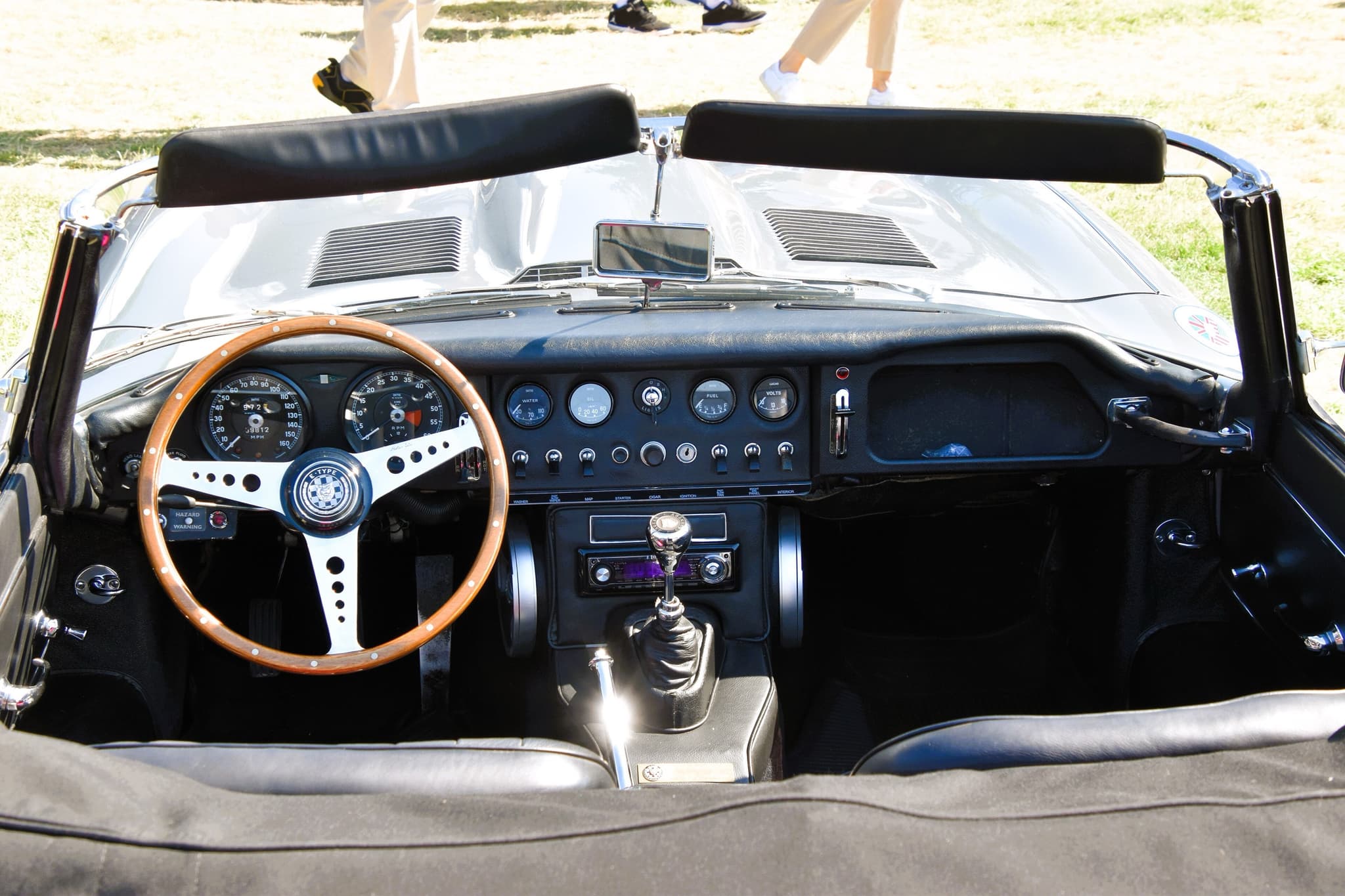 An E-Type roadster's cockpit seen from behind, with a black centre panel of dials and toggle switches and a wood-rim wheel on the left.