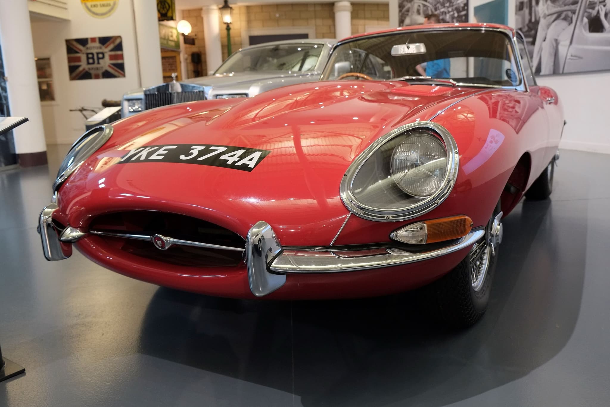 A red E-Type coupé with wire wheels in a museum hall, a silver Rolls-Royce behind it.