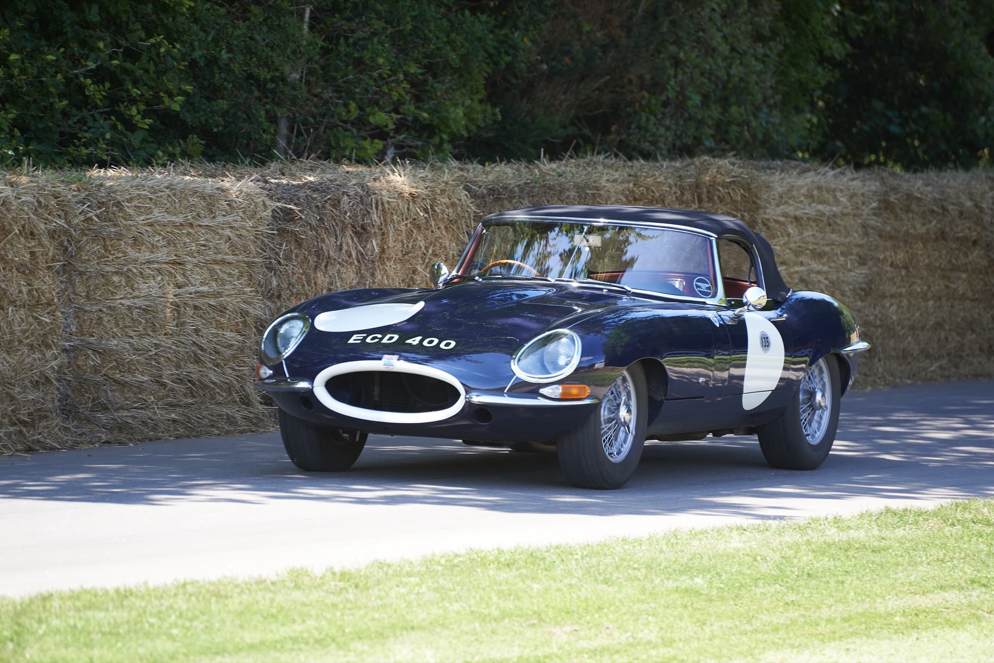 A dark blue E-Type roadster with a hardtop, white roundels and the registration ECD 400 on a road lined with straw bales.