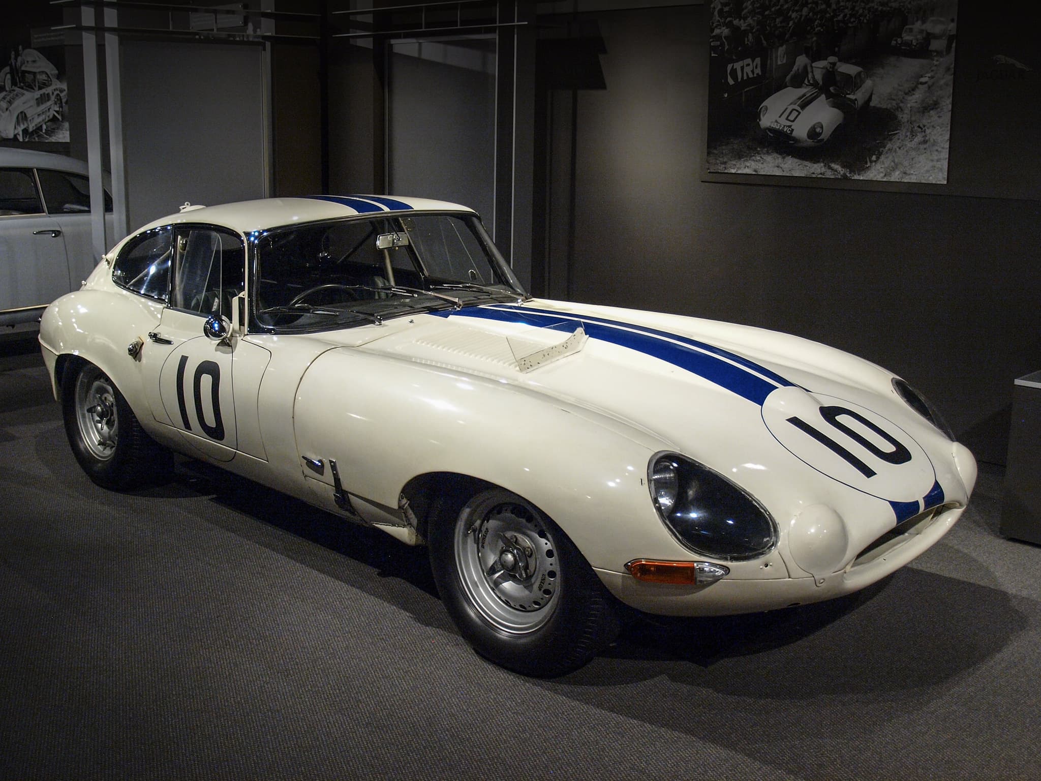 A white E-Type coupé with two blue stripes and the number 10 in a dimly lit museum. Opens a larger view