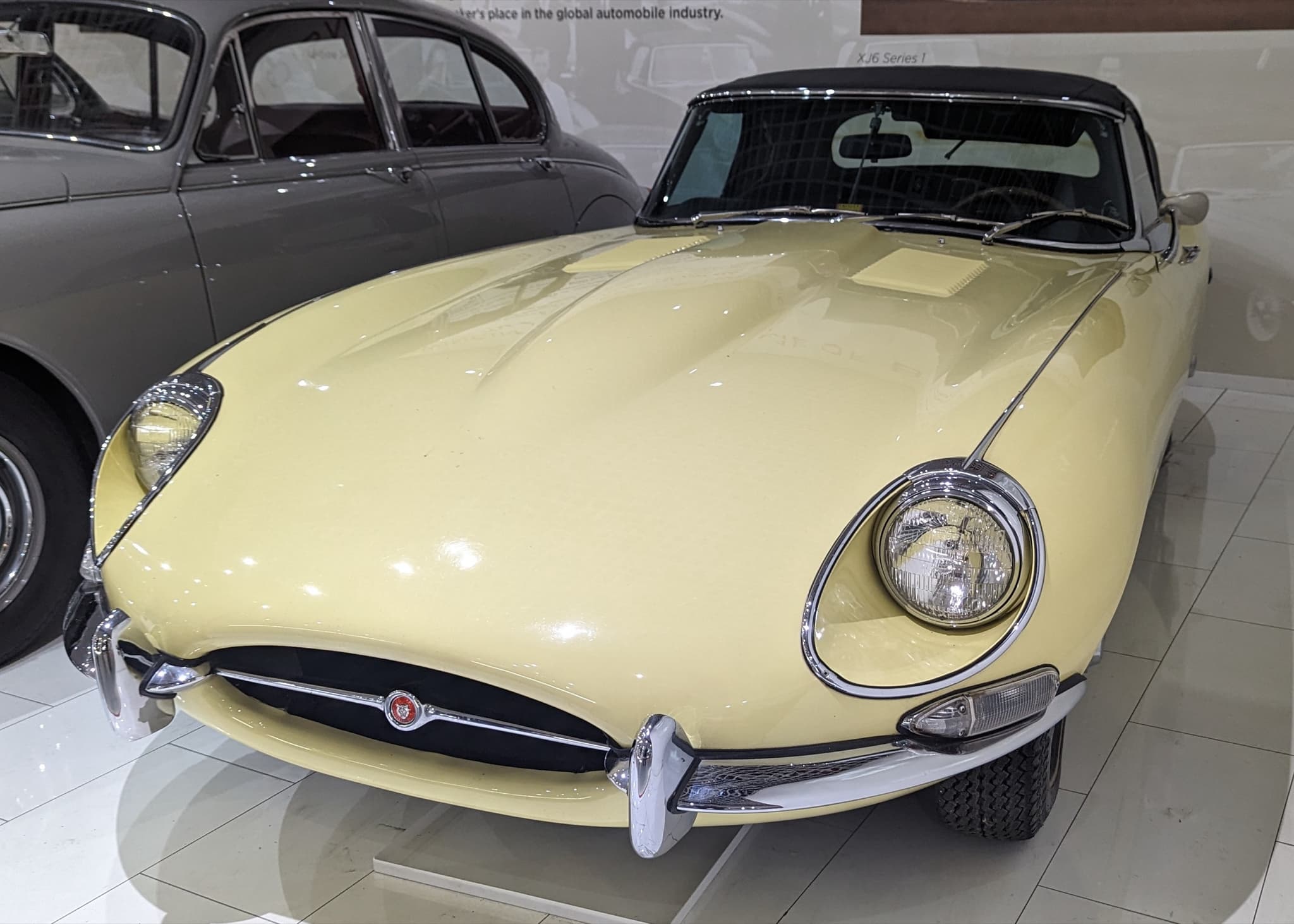 A pale yellow E-Type roadster with its black hood up and open headlamps, on white tiles in a museum.