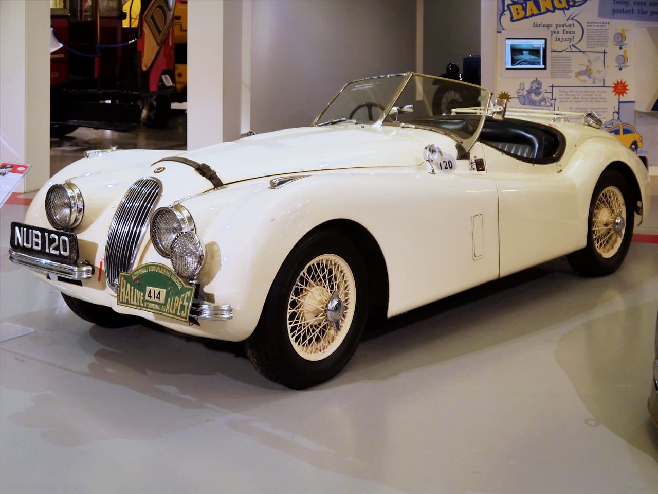 A white Jaguar XK120 roadster with the registration NUB 120 and a rally plate in a museum.
