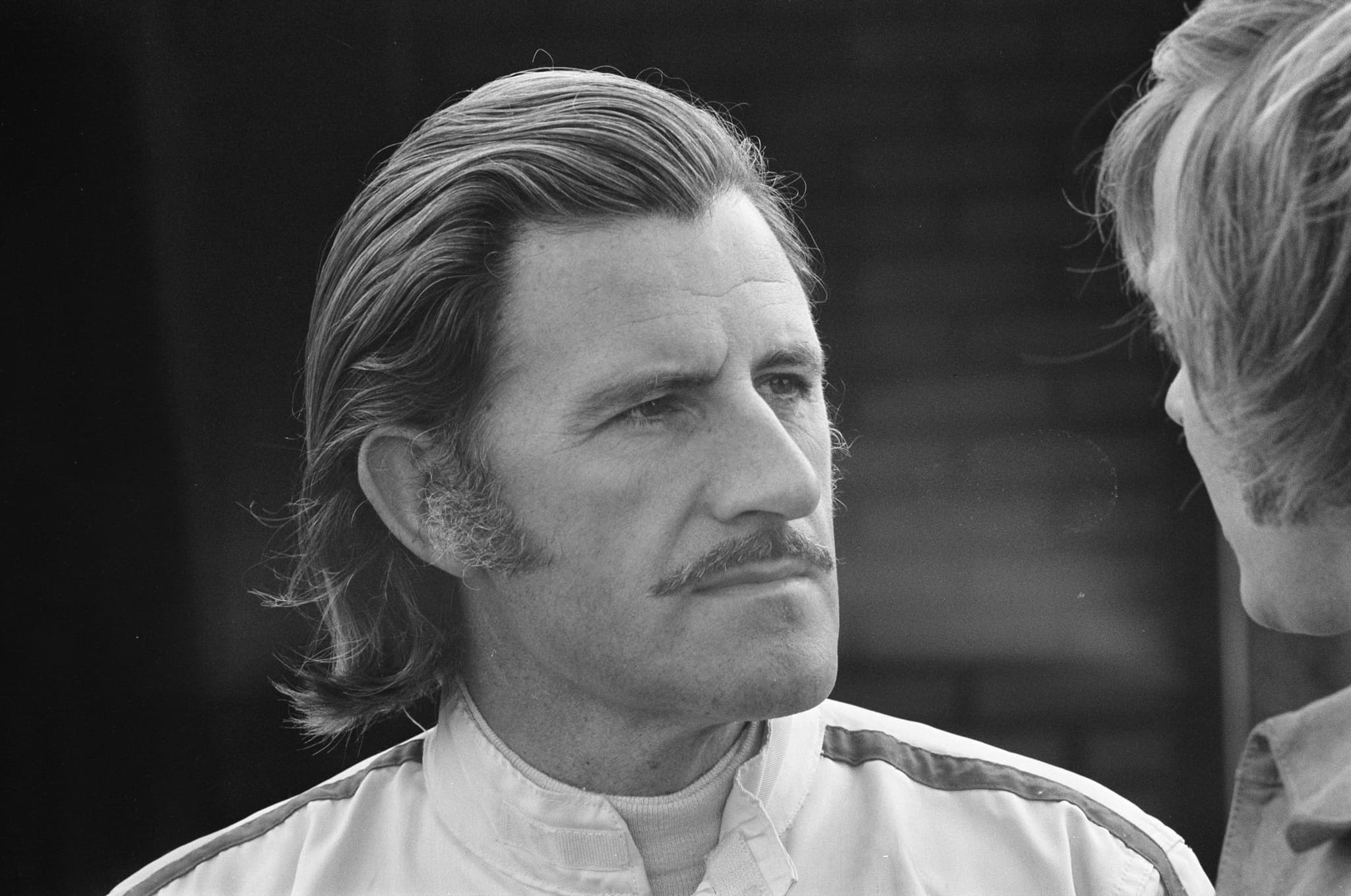 A black-and-white portrait of a man with swept-back hair and a moustache, wearing white racing overalls. Opens a larger view