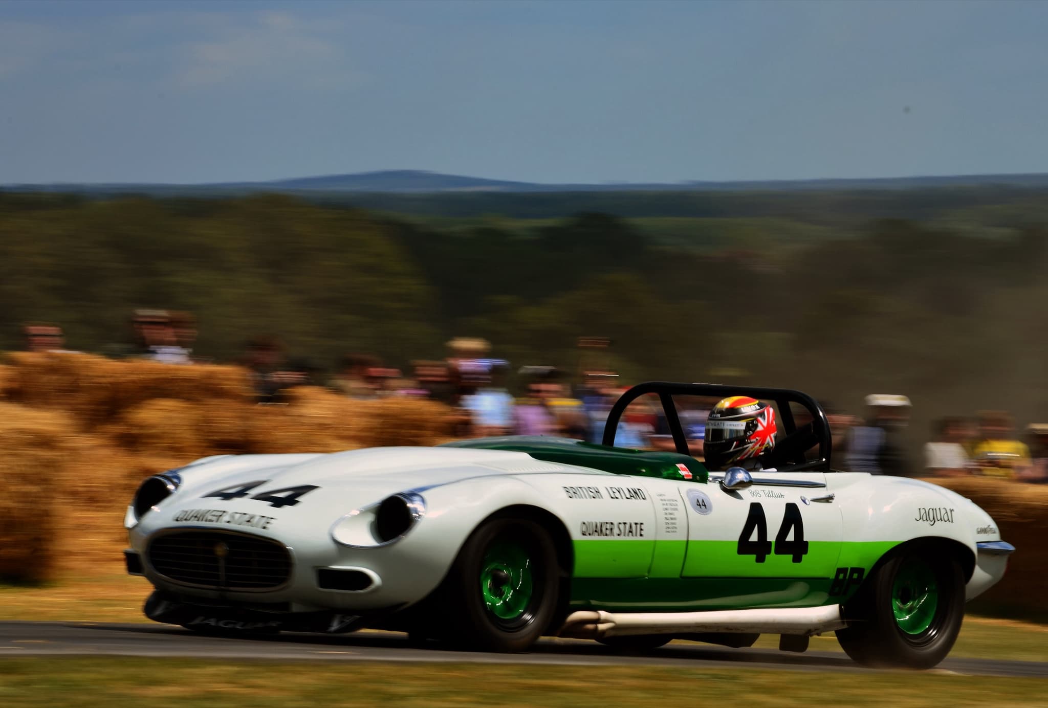 A white and green E-Type V12 race car numbered 44 driving fast past straw bales.