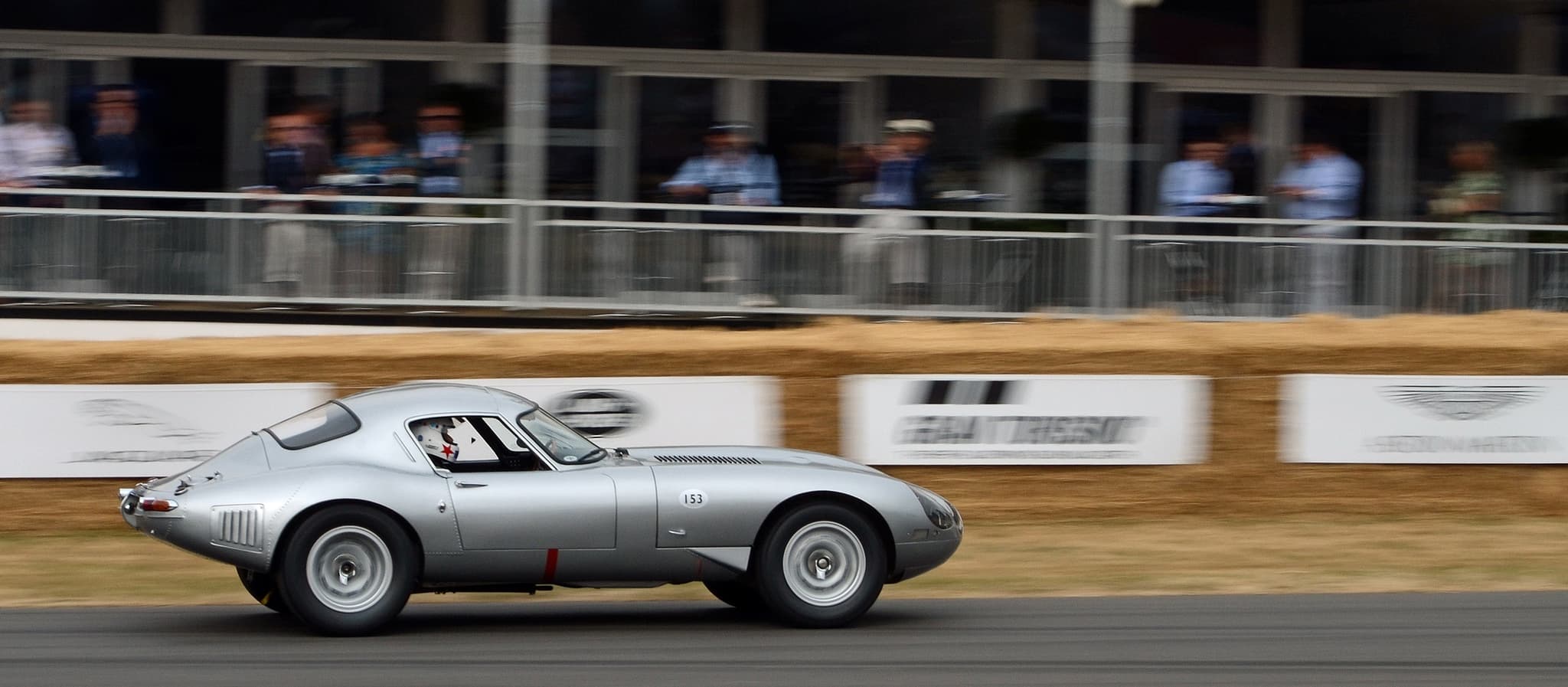 A silver low-drag E-Type coupé driving fast past straw bales and banners.