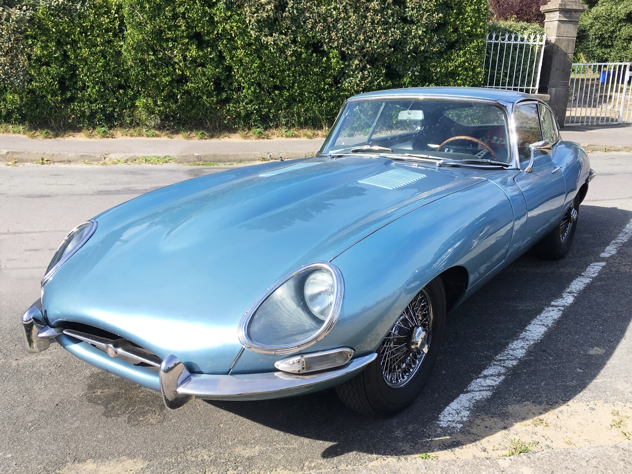 A pale blue E-Type with a fixed roof and worn paint, parked on a street beside a hedge and an iron gate.