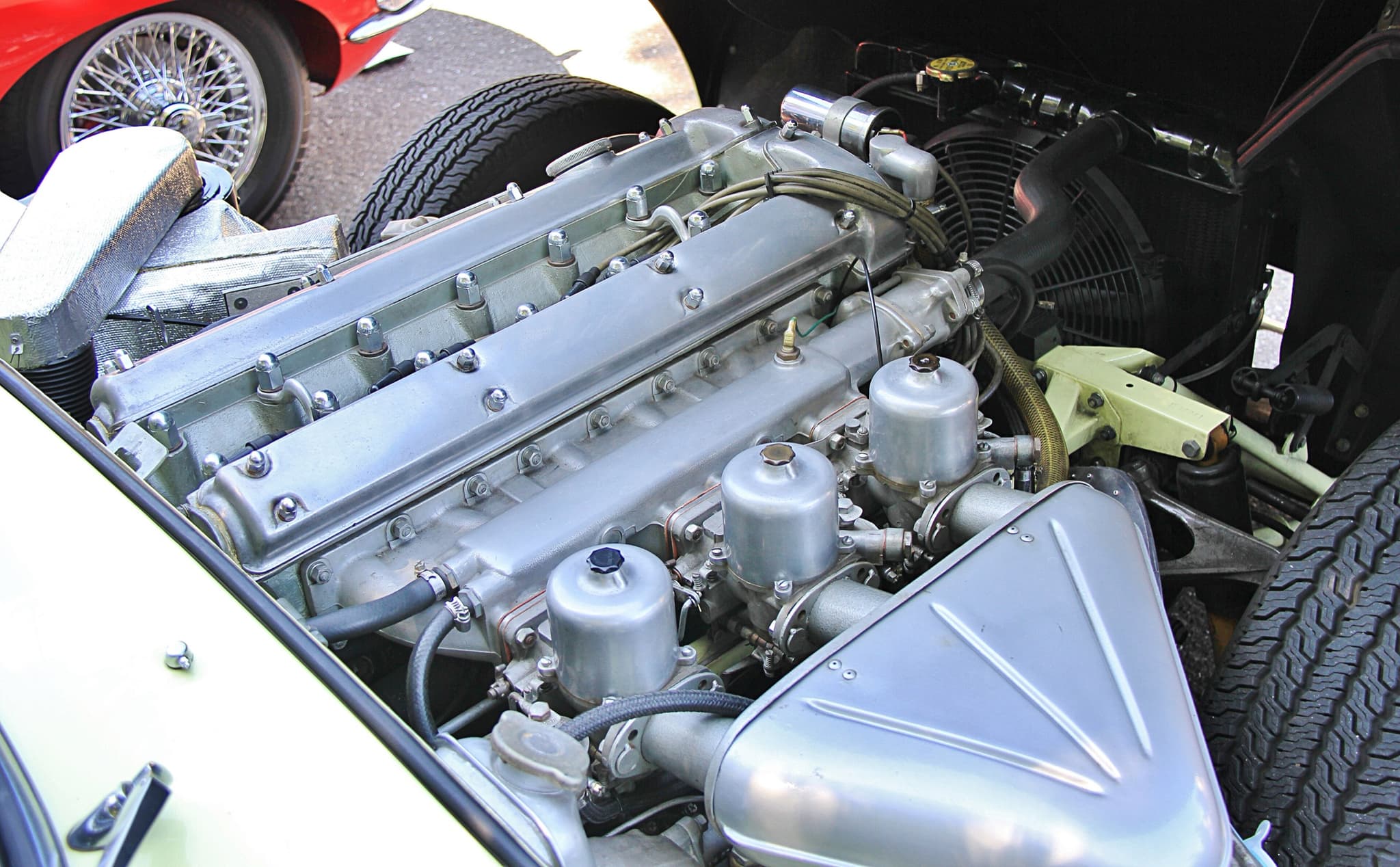An E-Type's six-cylinder engine with three carburettors under a ribbed aluminium air box, in a cream-painted engine bay.