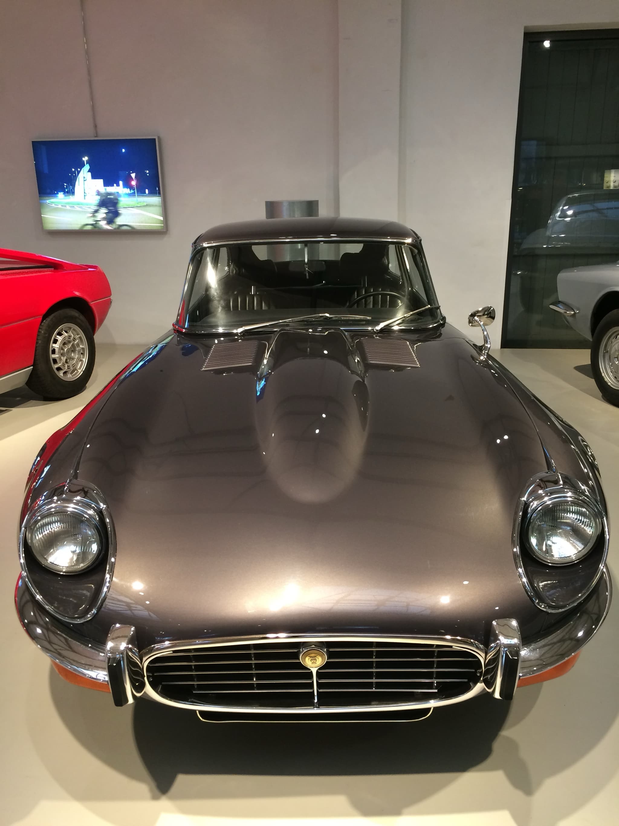 A grey E-Type 2+2 seen head-on in a museum, showing its cross-hatched grille and bulged bonnet.