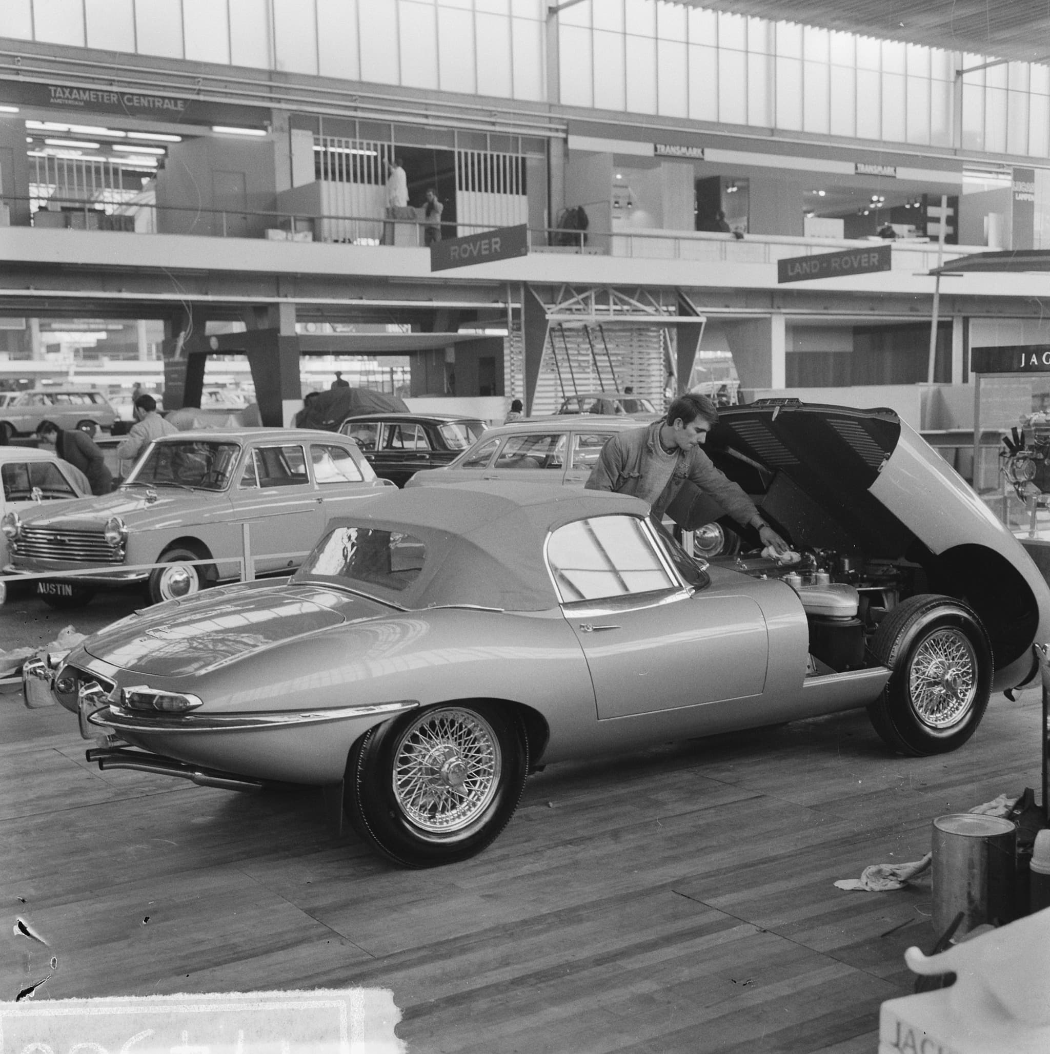 Black-and-white photograph of a motor show being set up, with an E-Type roadster on a stand and a worker at its open bonnet.