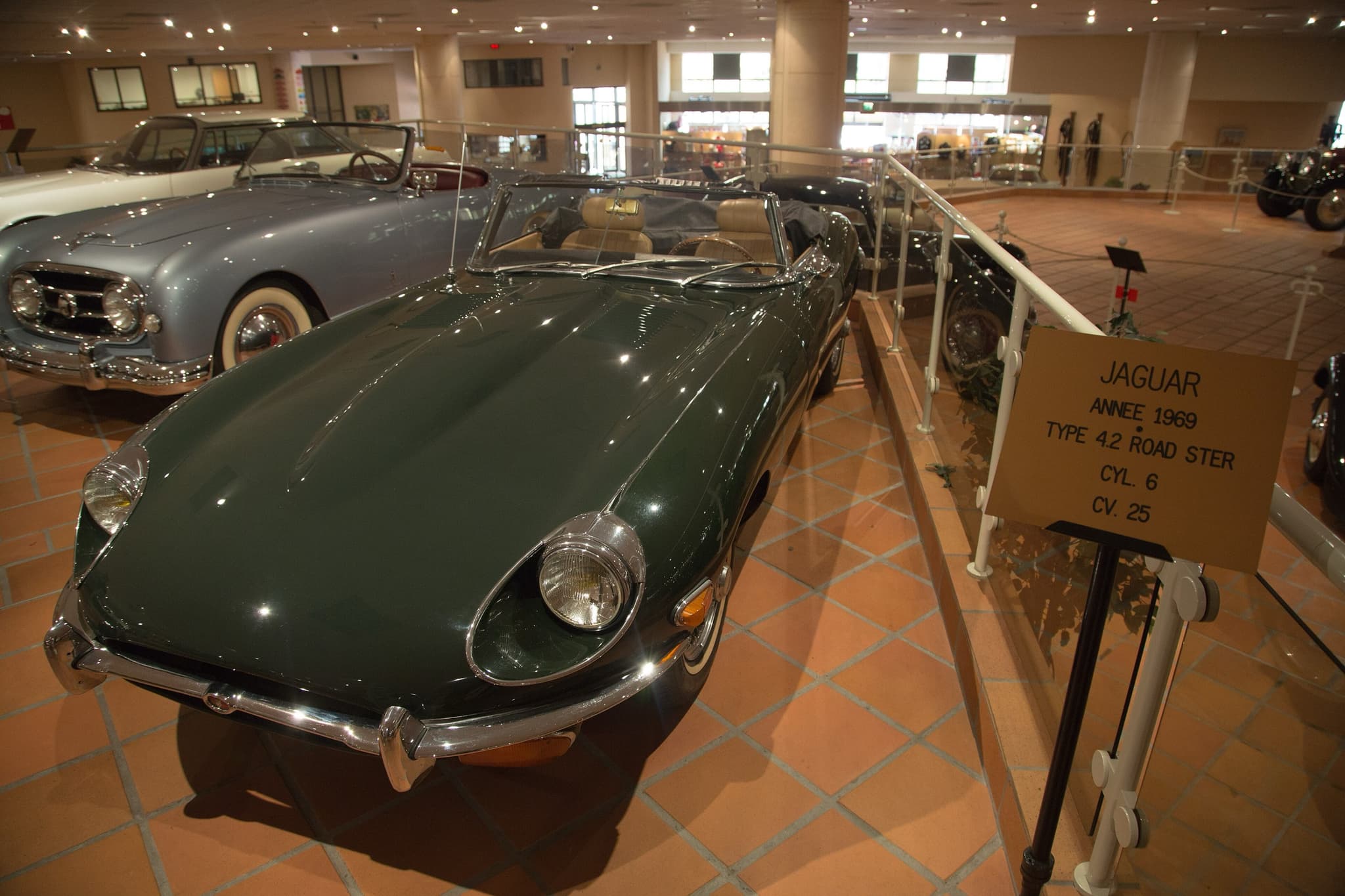 A dark green E-Type roadster with open headlamps and a large oval intake on terracotta tiles in a museum. Opens a larger view