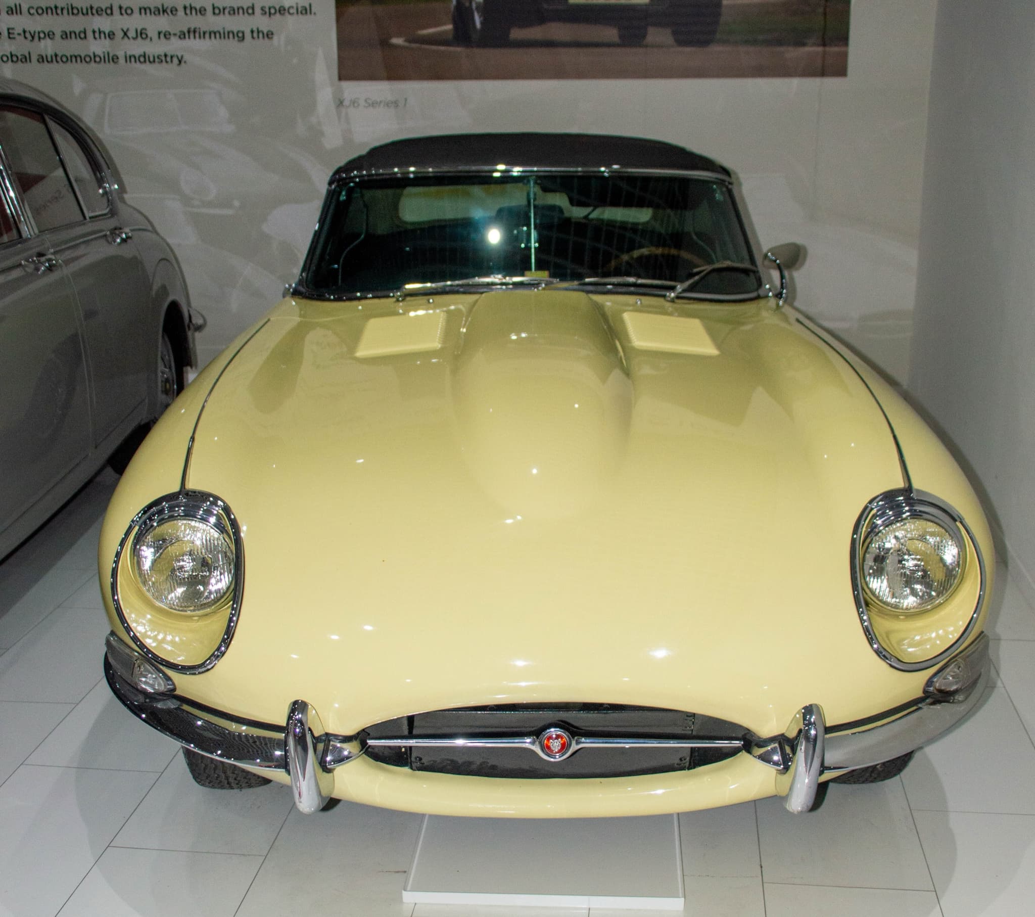 A pale yellow E-Type roadster with its hood up seen from above the nose, showing open headlamps and a small oval intake.