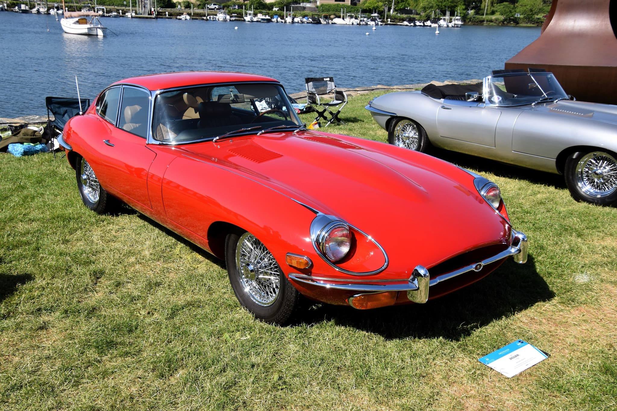 A red E-Type coupé on grass beside a harbour, with a large oval intake and the indicators below the bumper.